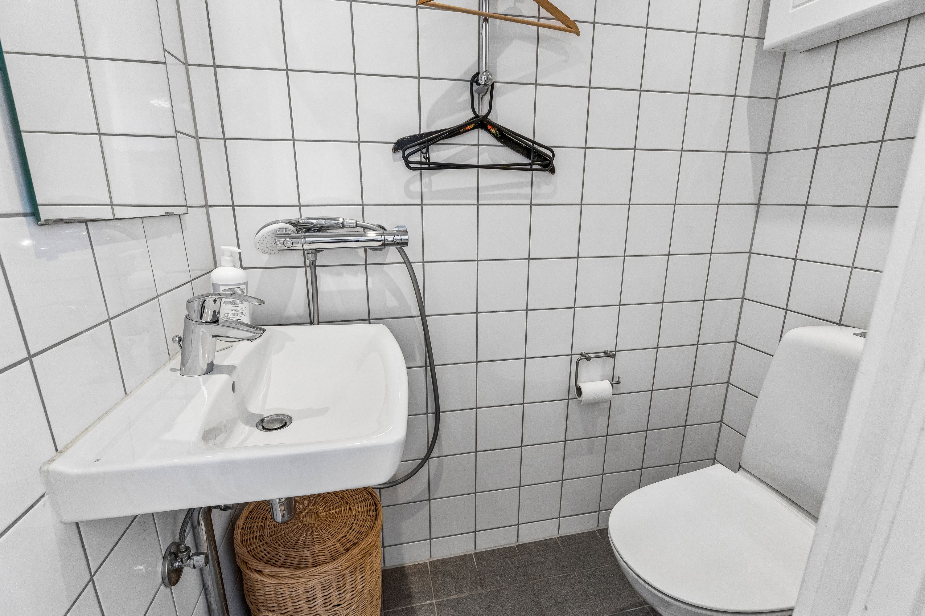 Et hvidt toilet med håndklædeholder, gulvvarme og en lille hængeklædeskabs på hjørnet i et lille badeværelse med hvidt flisebelægning og hvidt toilet