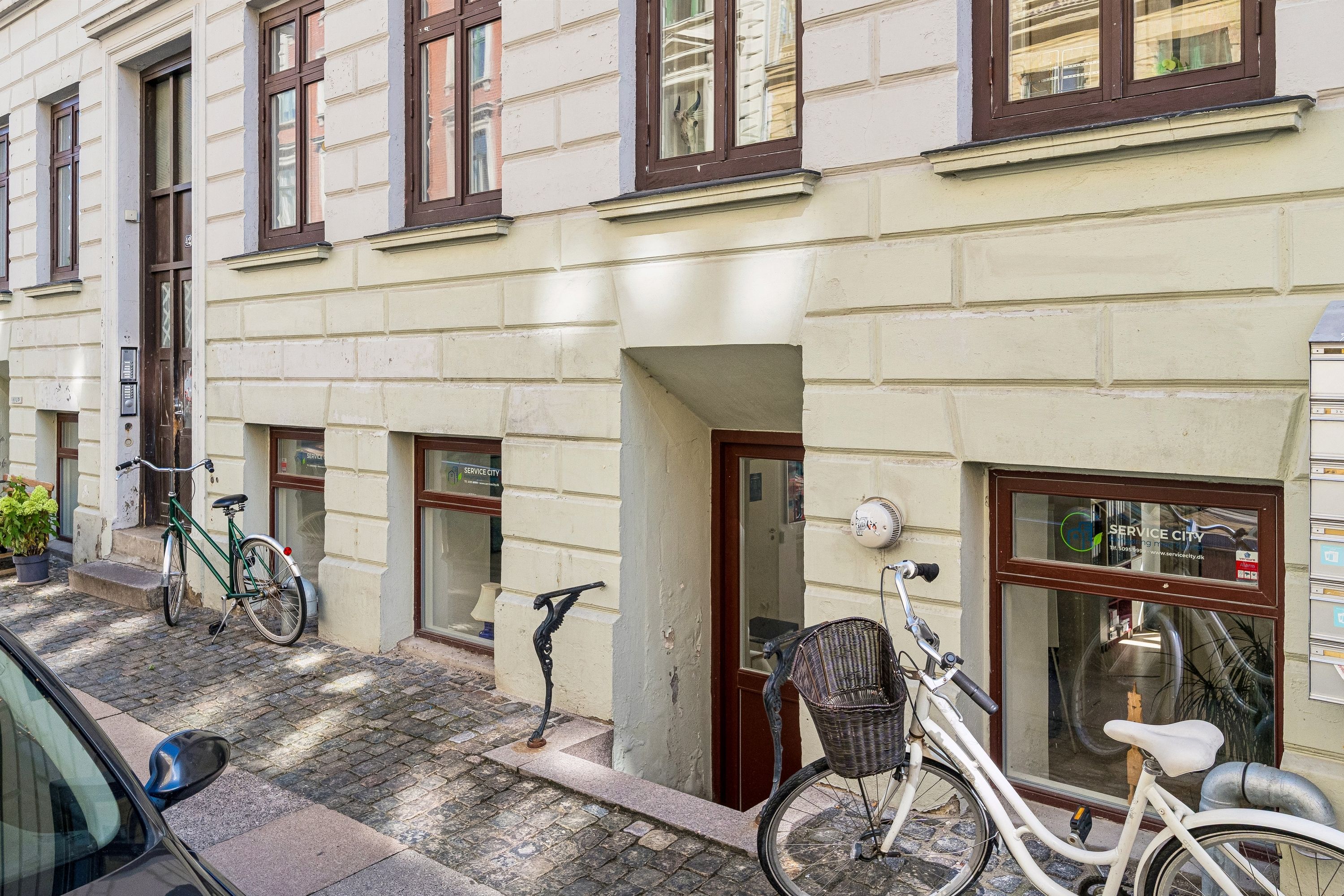 Butik til leje på Peter Fabers Gade 42 med to parkerede cykler og en hvid bil parkeret på siden af gaden. Service City skrift på vinduerne og en postkasse på hjørnet