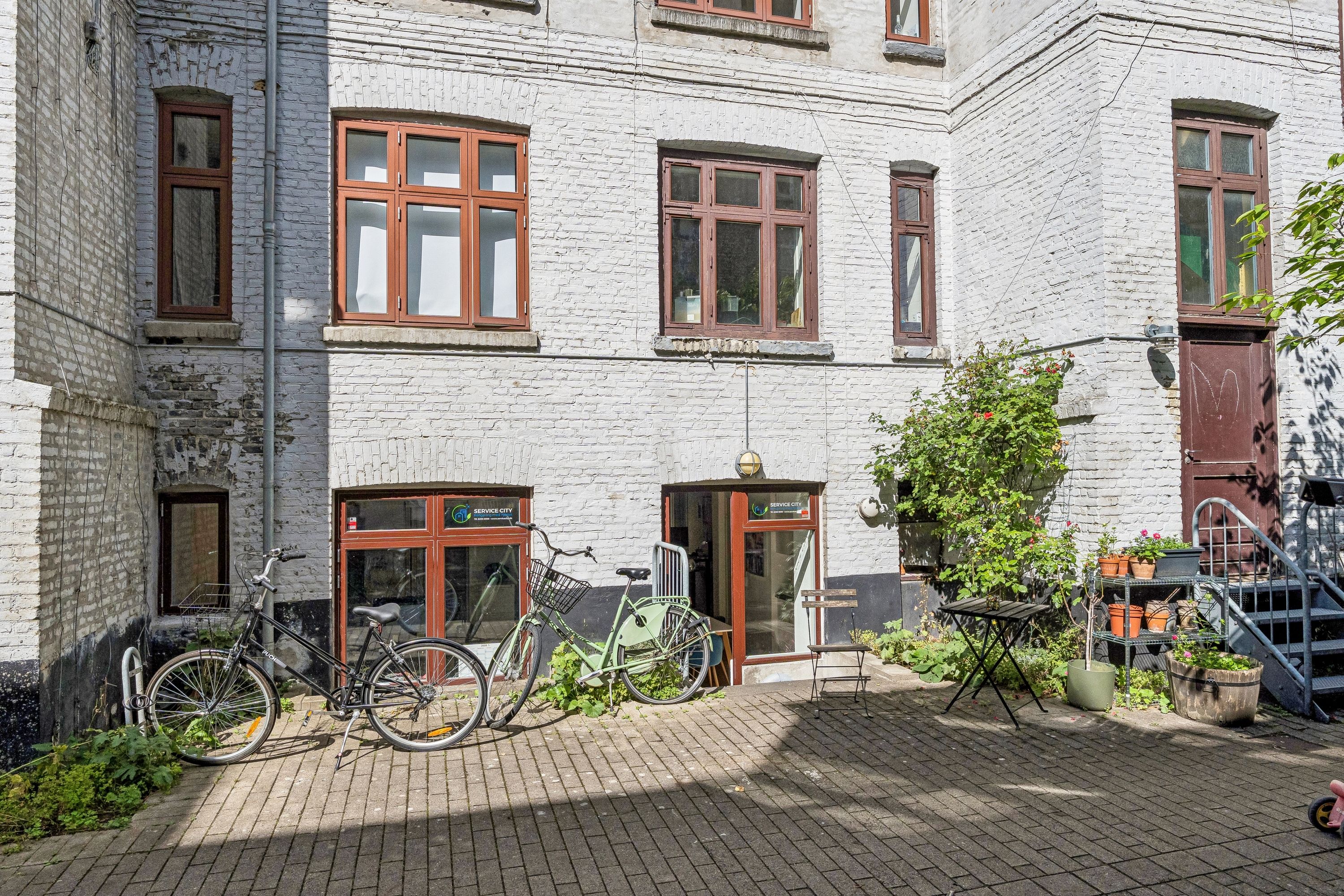 Lejet butik i høj kælder med vinduer og blomster i haven og to cykler parkeret udenfor.
