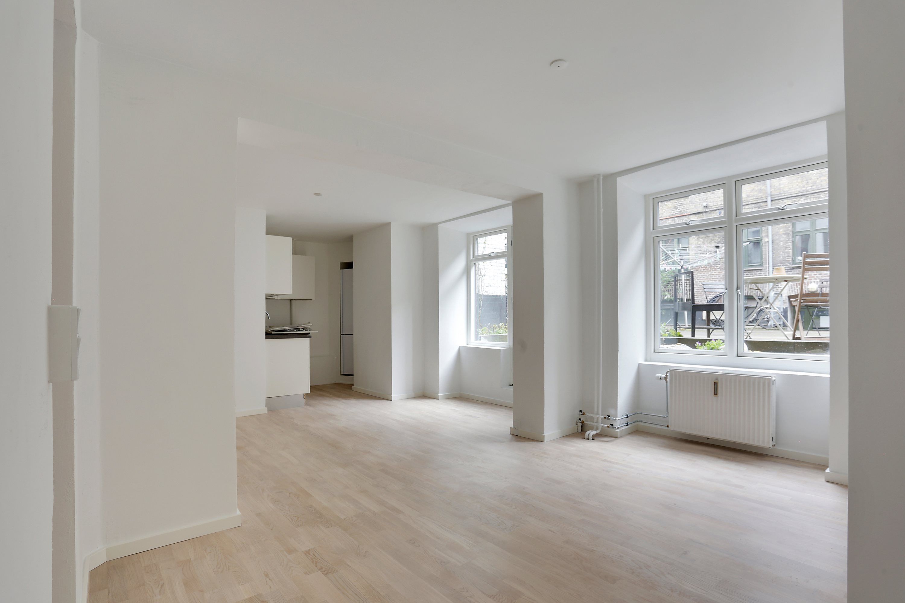 Store hvide vægge og lysegule trægulve i højkælderlejlighed. Solrige vinduer med udgang til altan med spisestedet og hængekøkken. Vægmonter i hjørnet og radiator nederst i væggen