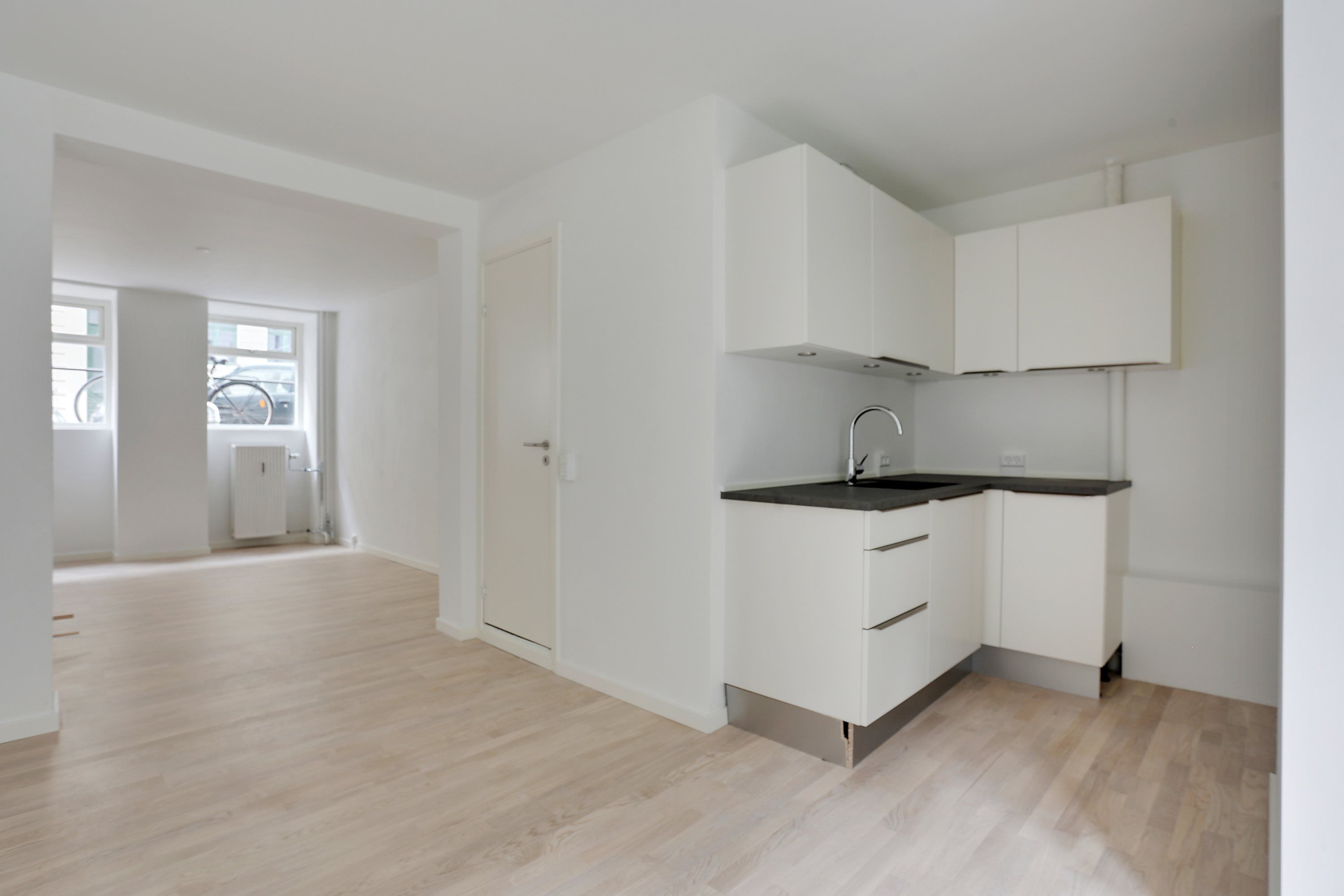 Hvidt køkken med lyse trægulv og hvide skabe, lysekroner og høj kvalitet. Slank vaskebækken med vandhane og hvidt loft. Vindue med udsigt til gaden.