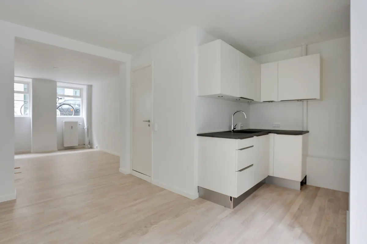 Hvidt køkken med lyse trægulv og hvide skabe, lysekroner og høj kvalitet. Slank vaskebækken med vandhane og hvidt loft. Vindue med udsigt til gaden.