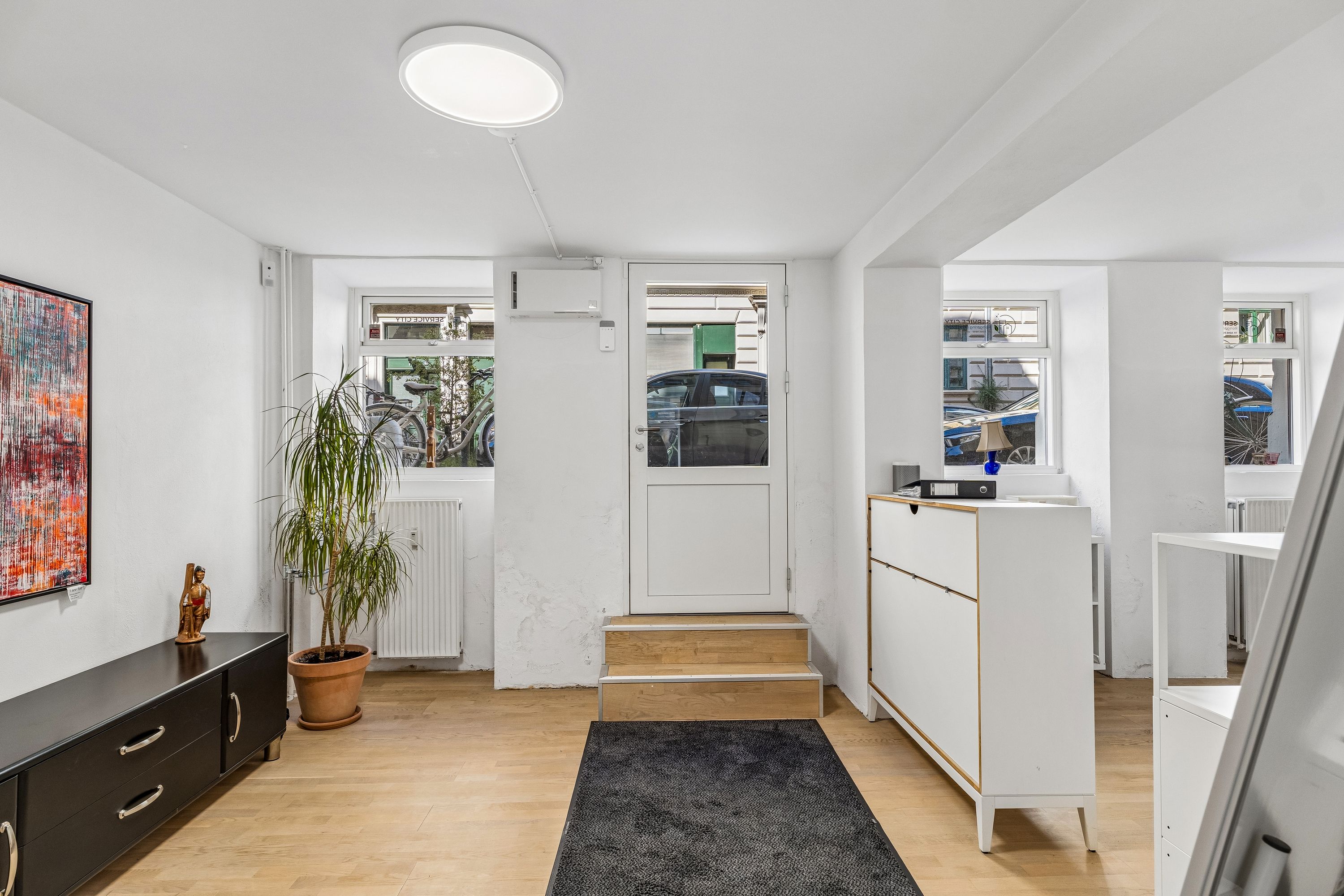 Indvendigt i en renoveret kælderlejlighed på Peter Fabers Gade. Lejligheden har hvidt interiør med en plante, kunstværk og et hvidt sidebord. En hvid dør og vinduer med udsigt til en bilparkering.