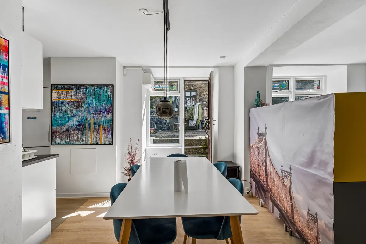 Moderne stue med hvidt trægulv og lyse vægge, møbler og kunstværk. Værelset har en lang hvid spisebord og lyse blå stole. Billedet viser lyse lysekroner og åben dør til gård.