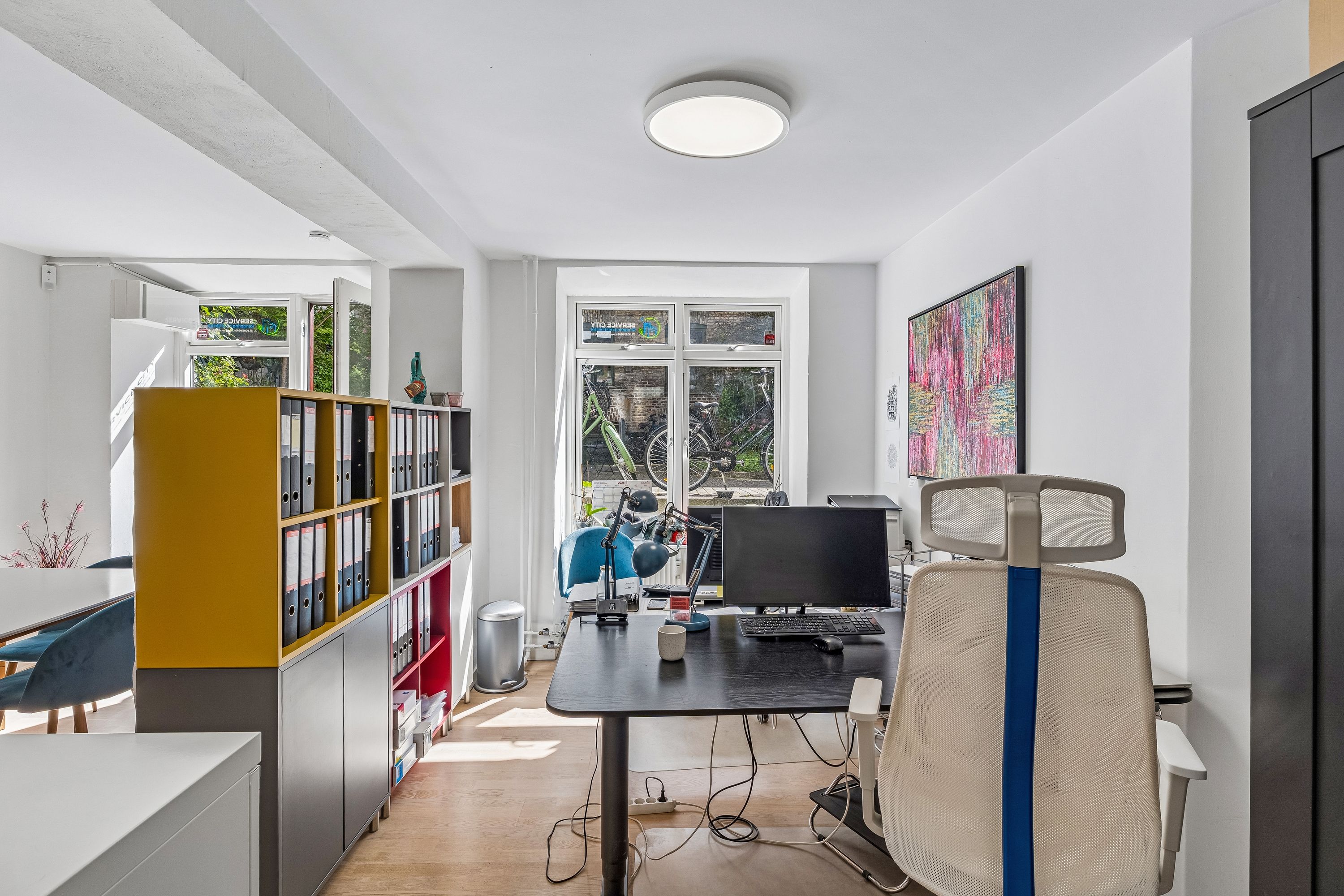 Billede af lejlighed med et moderne kontor, hvor der er et skrivebord med computer og stol samt en hylde med bøger og filer