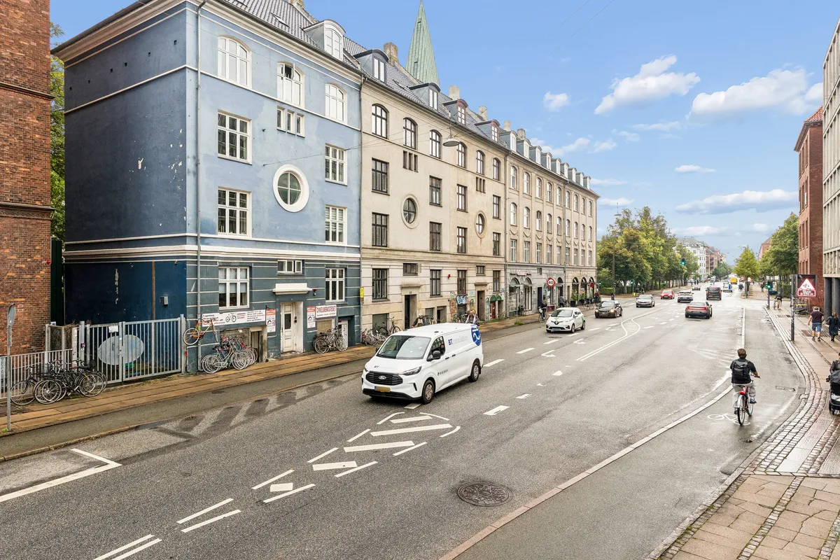 Forskudt hus med høj kælderlejlighed til leje på Enghavevej 51. Biler kører på to sporet gade. På fodgængerbroen er der folk, og en mand cykler.