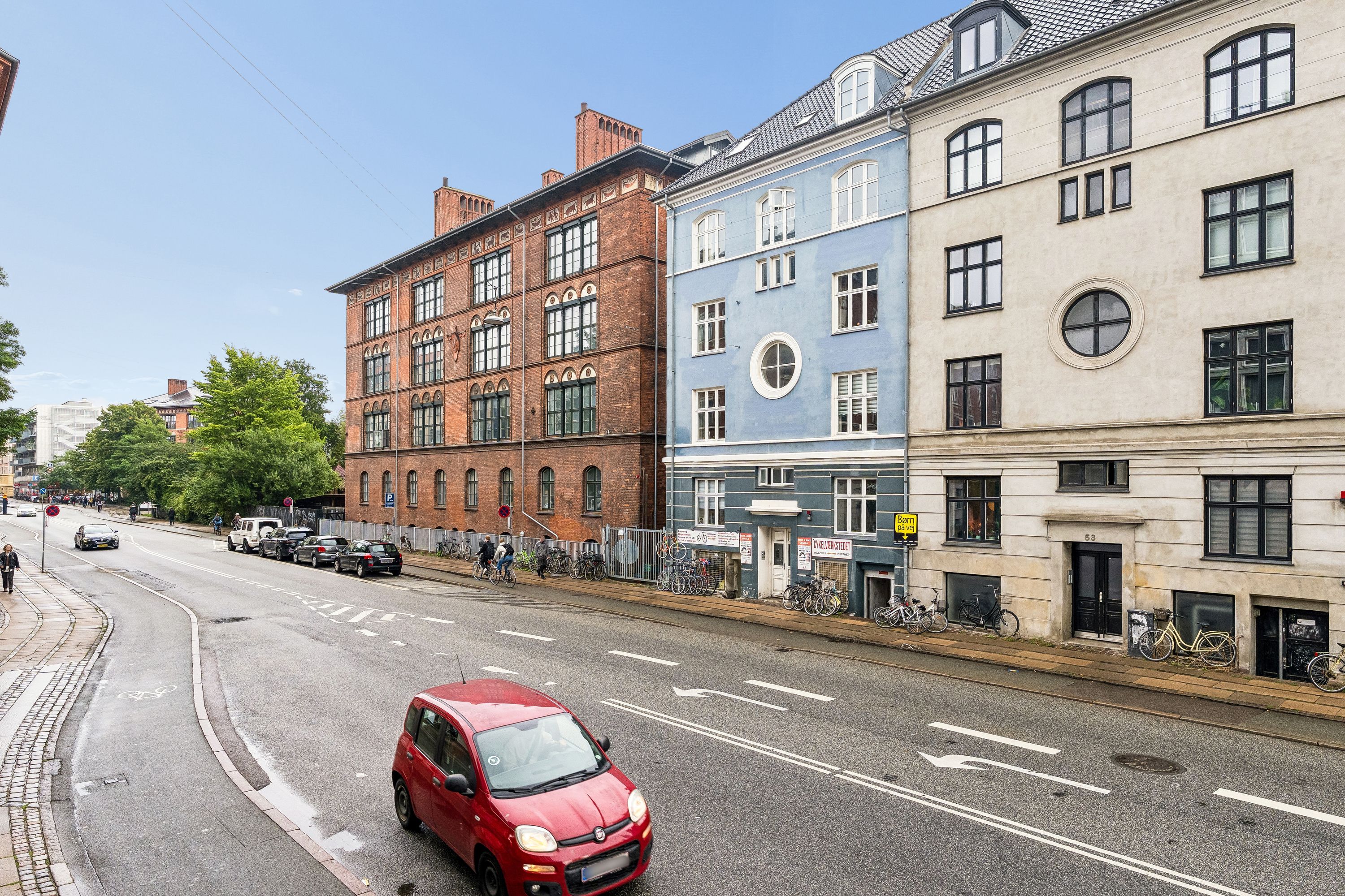 Red bil kører ad hovedgade med grønne træer og bygninger i baggrunden, med to store butikker og cykelparkering