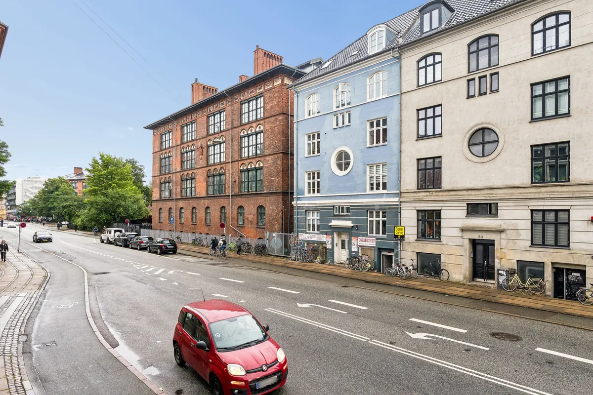 Red bil kører ad hovedgade med grønne træer og bygninger i baggrunden, med to store butikker og cykelparkering