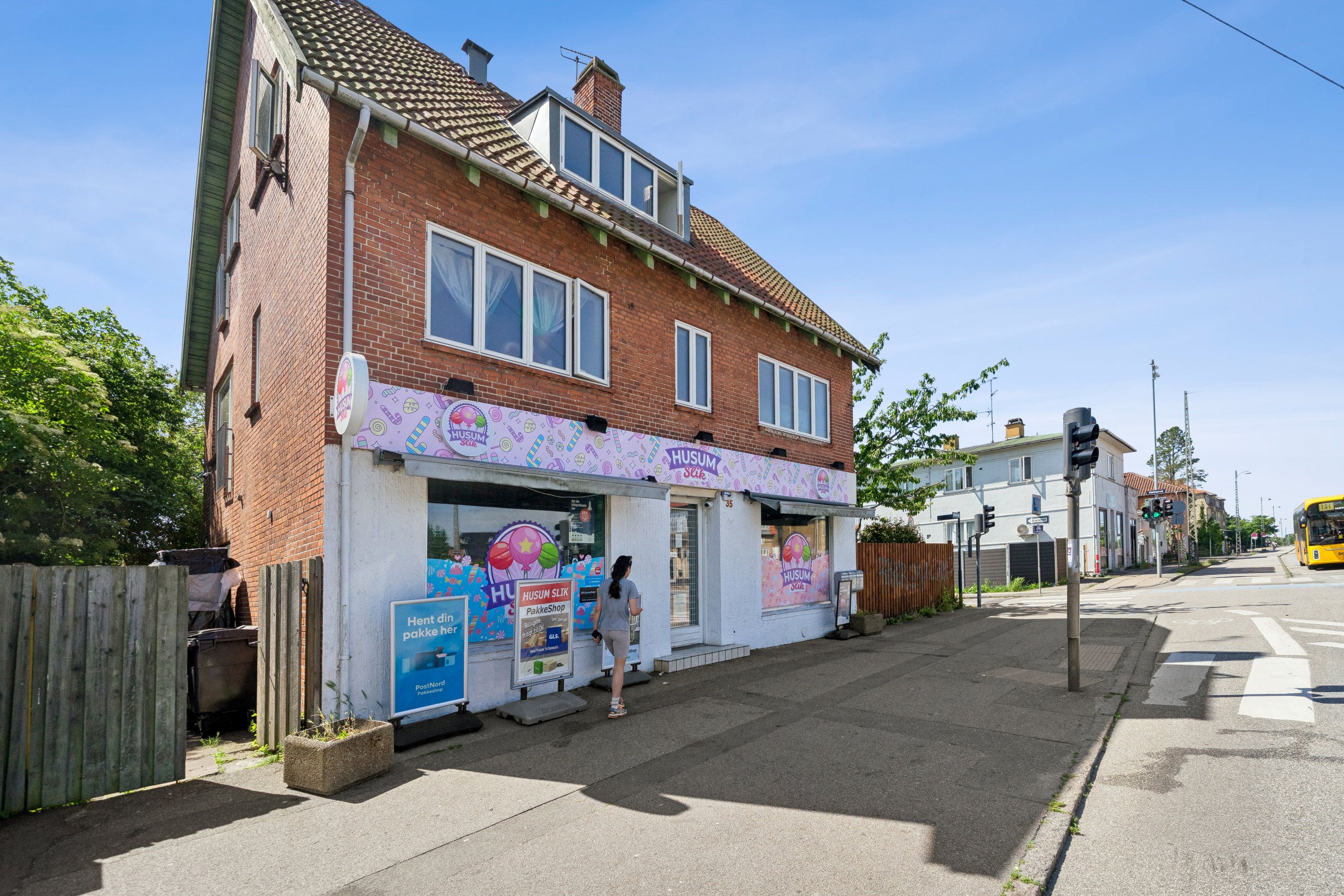En kvindelig gående forbi en butik med navnet Husum Slik, der sælger slik. Der er også et skilt med teksten Pakkehop, og en bus kører forbi på den anden side af gaden