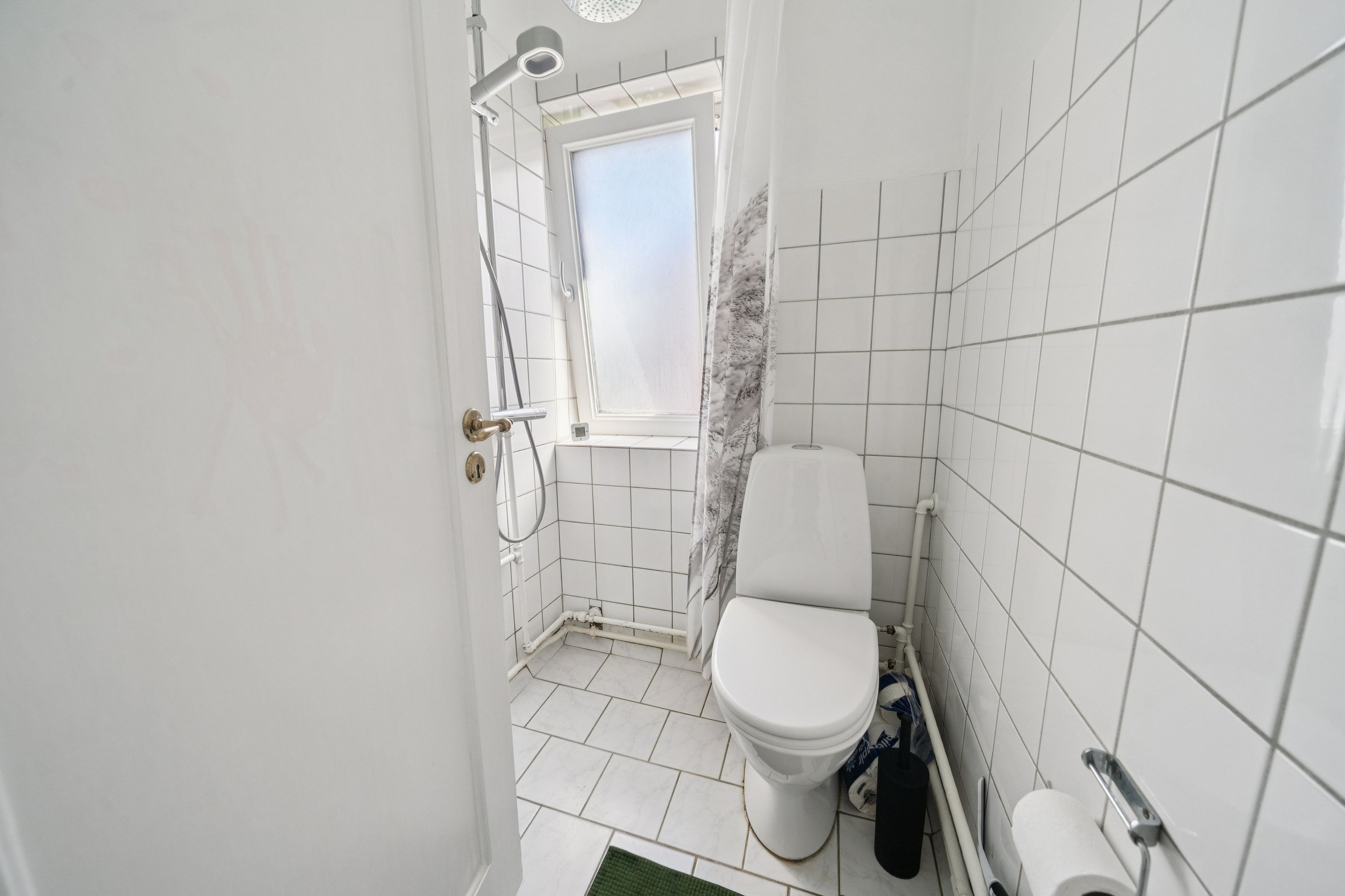 En ren, hvidt fliserne badeværelse med en toilet, en bruser, et toiletpapir og et tørklæde i det lille vindue