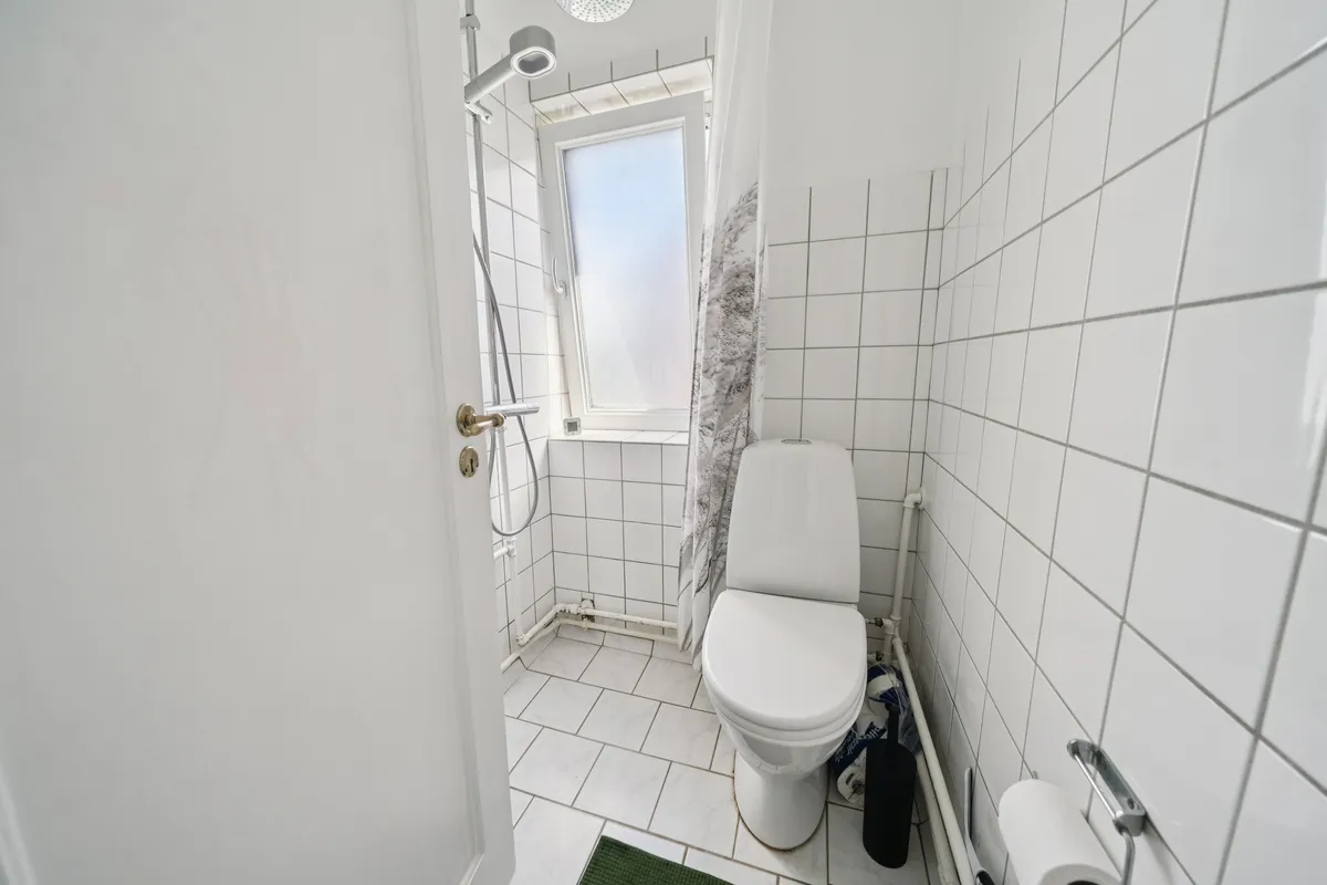 En ren, hvidt fliserne badeværelse med en toilet, en bruser, et toiletpapir og et tørklæde i det lille vindue