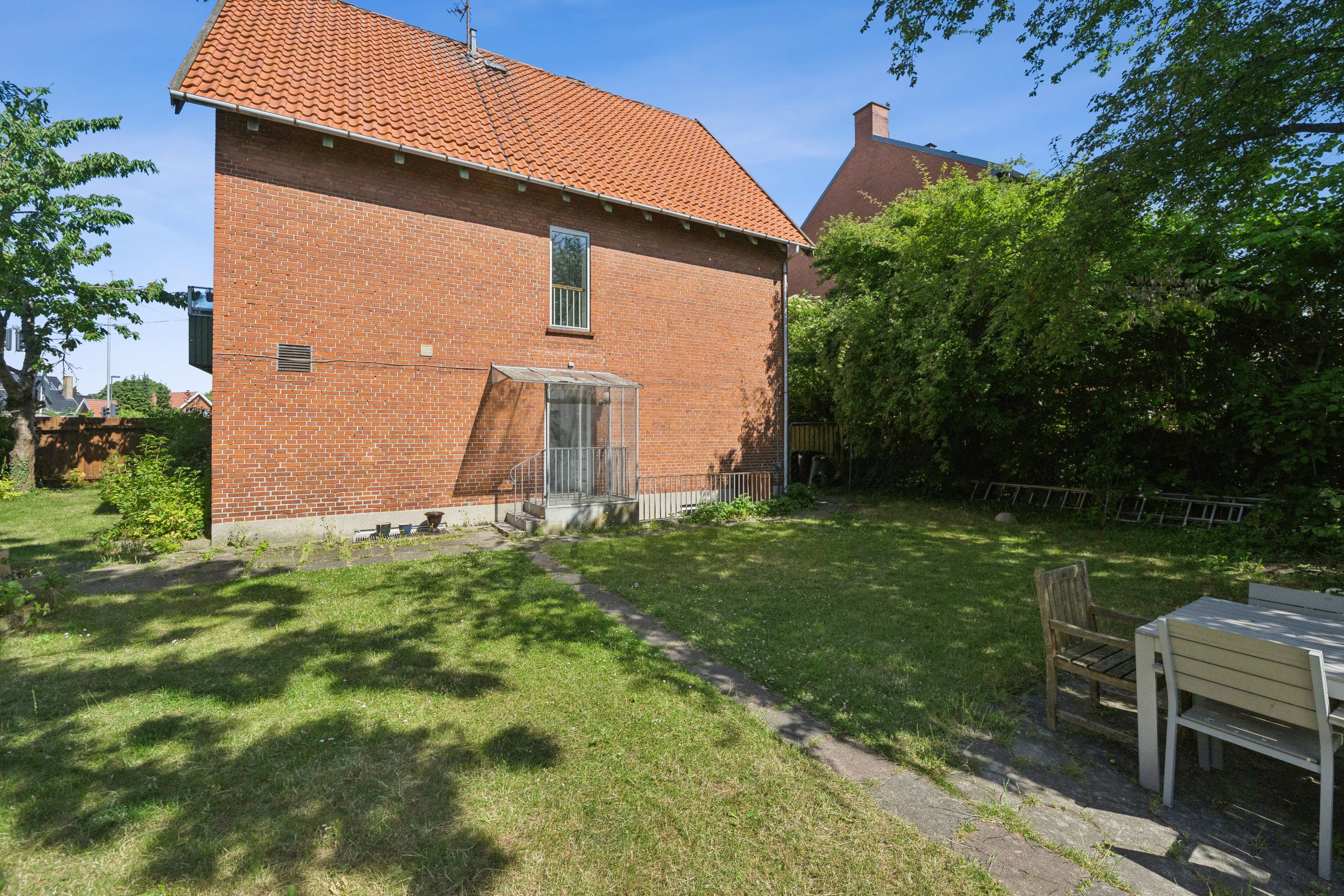 En rækkehus med rød mursten og grønne græsplæner, der er omgivet af træer og planter. Et bord og stol er placeret på terrassen, der vender mod det lyse himmelblå.