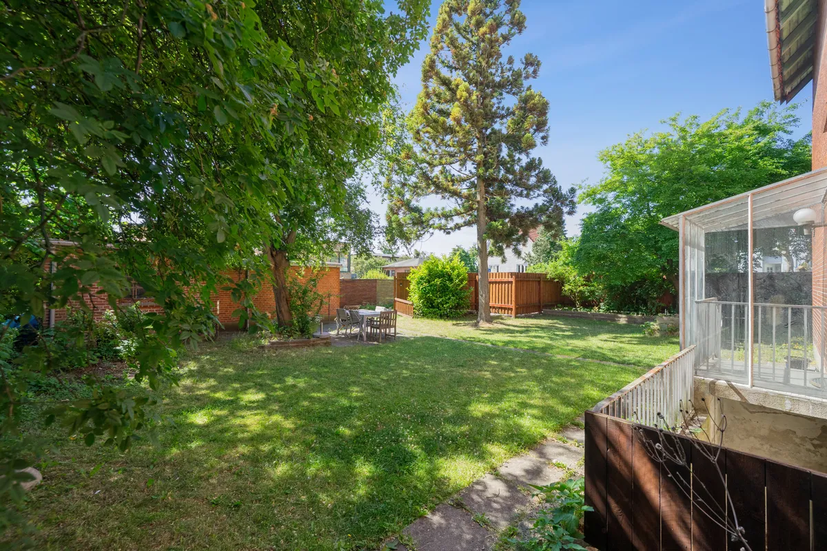 En stor have med græsplæne og træer samt en terrasse med udendørsbord og stole. En række røde murstensbygninger og en lys dag.