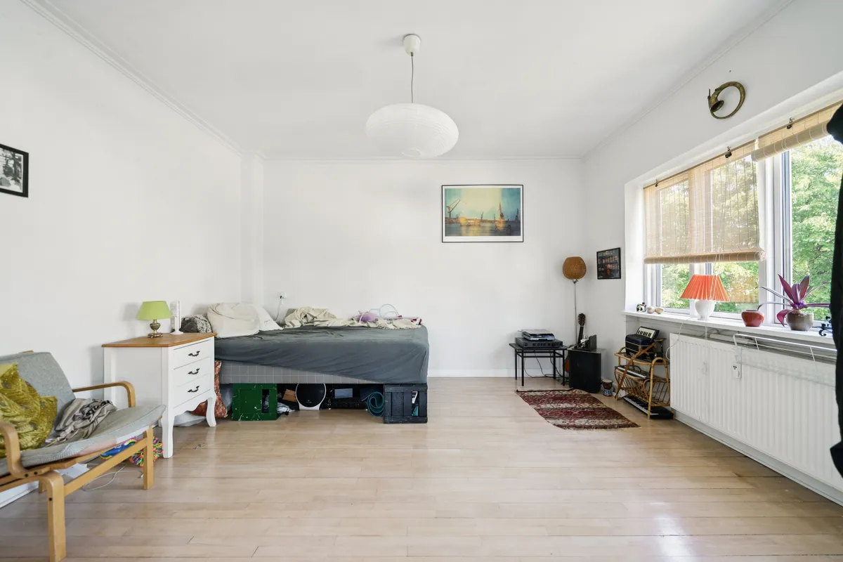 Et enkeltværelse med seng, trægulv, hvidt køkken, kommode, puder, lampe, kunstværk og et vinduesvisir med plante og radiator