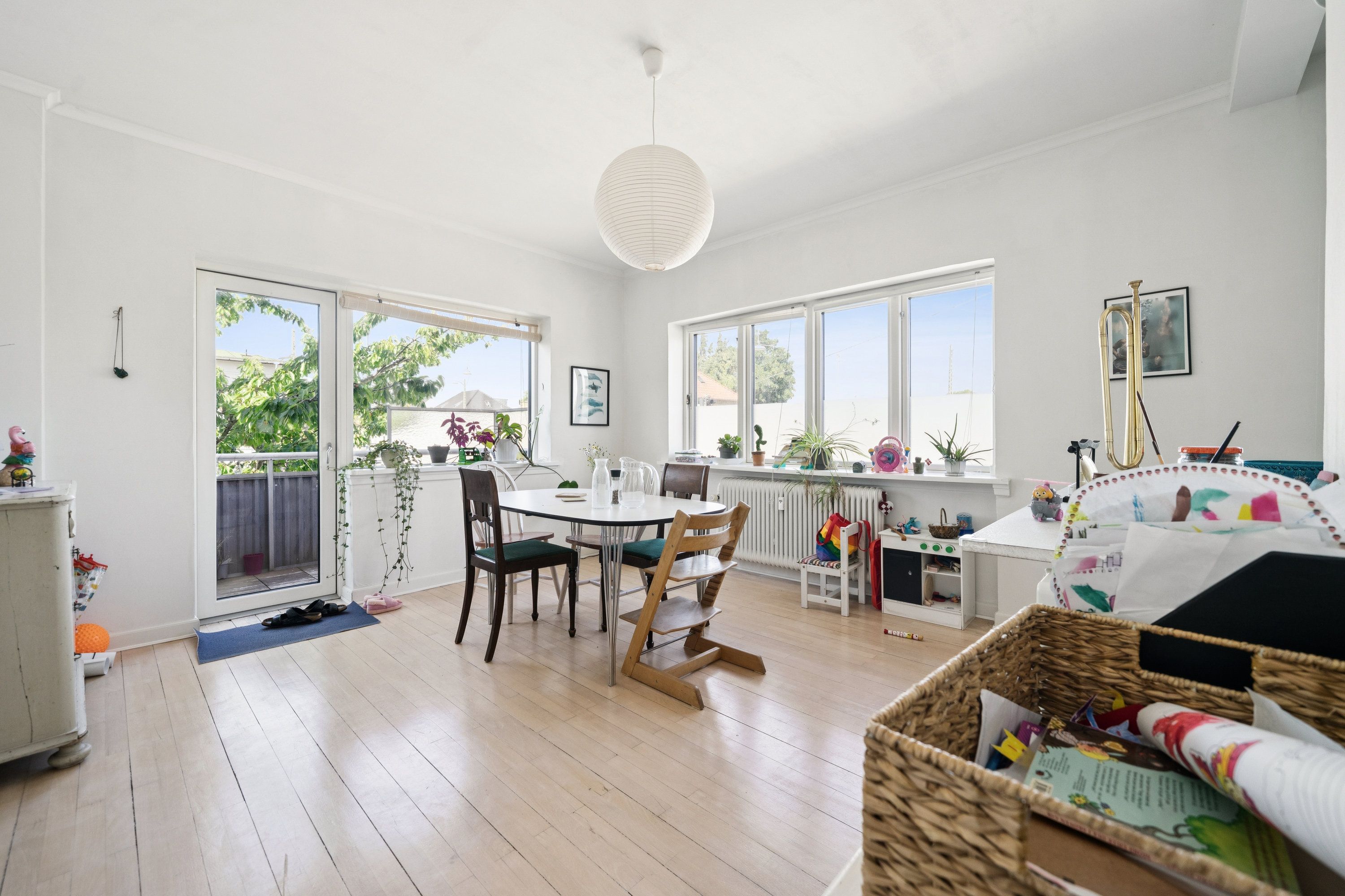 Værelse med hvidt loft og vægge, bord, stole, trægulv og vinduer med udgang til balkon. Planter og træer bagved vinduet og tøj i kurv og på sofa