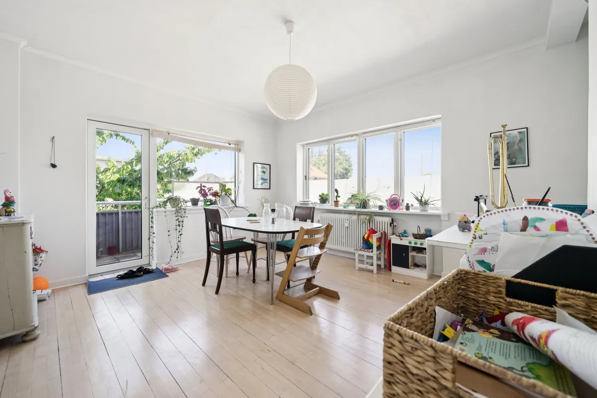 Værelse med hvidt loft og vægge, bord, stole, trægulv og vinduer med udgang til balkon. Planter og træer bagved vinduet og tøj i kurv og på sofa