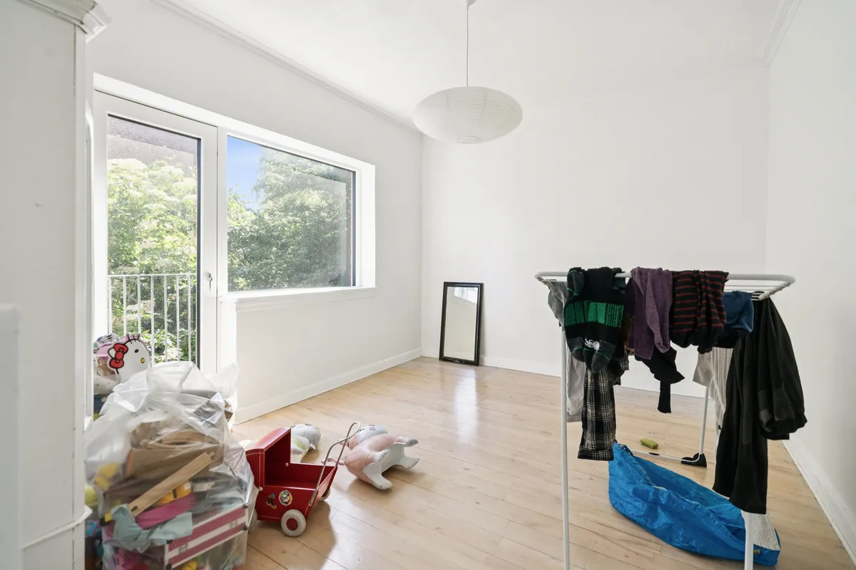 Værelse med lyse trægulve og hvide vægge, tøjstativ med tøj, legetøj og kæledyrsfigurer, fuldvindue med udsigt til grønne træer og bygninger, hvid lampe og spejl på væggen