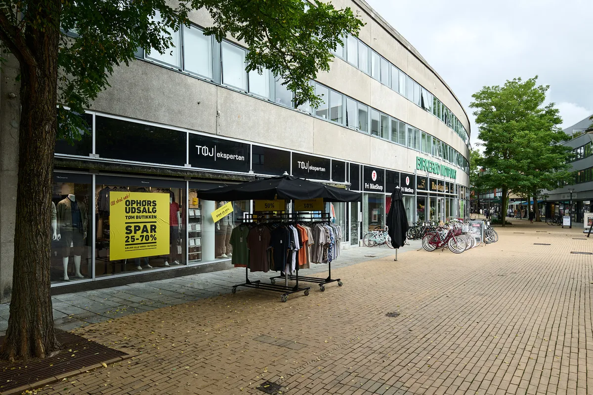 Butikslejemål med solbeskyttelse, tøj på hylder og mænd i vinduesvisning, samt cykler og træer bagved.