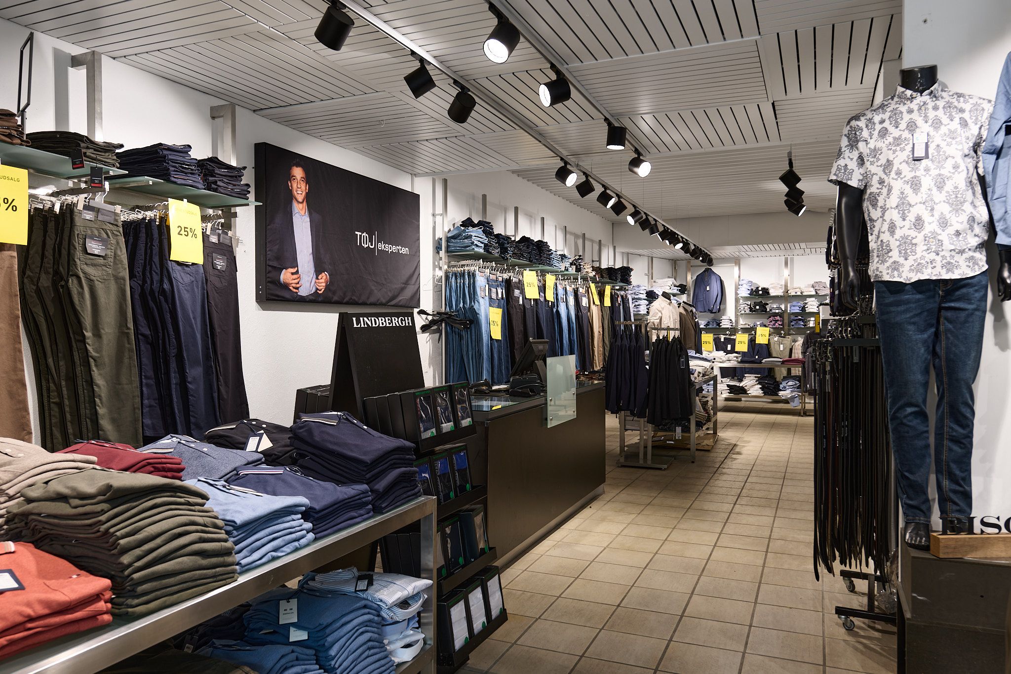 Butik med tøj, jeans og skjorter i et eksklusivt mærke, Lindberg. Prisskilte med 25% rabat, og mænd i blazer på væggen.