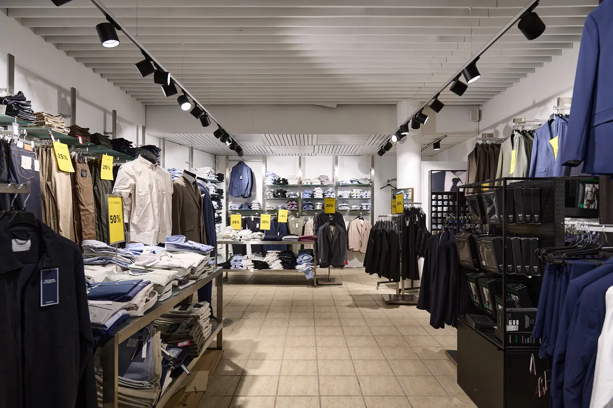 En butik med tøj og sko i stueplan. Der er tøj på hylder og på hængere. Der er udsalgsskilte på tøjet. Gulvet er fliselagt og der er lysekroner på loftet