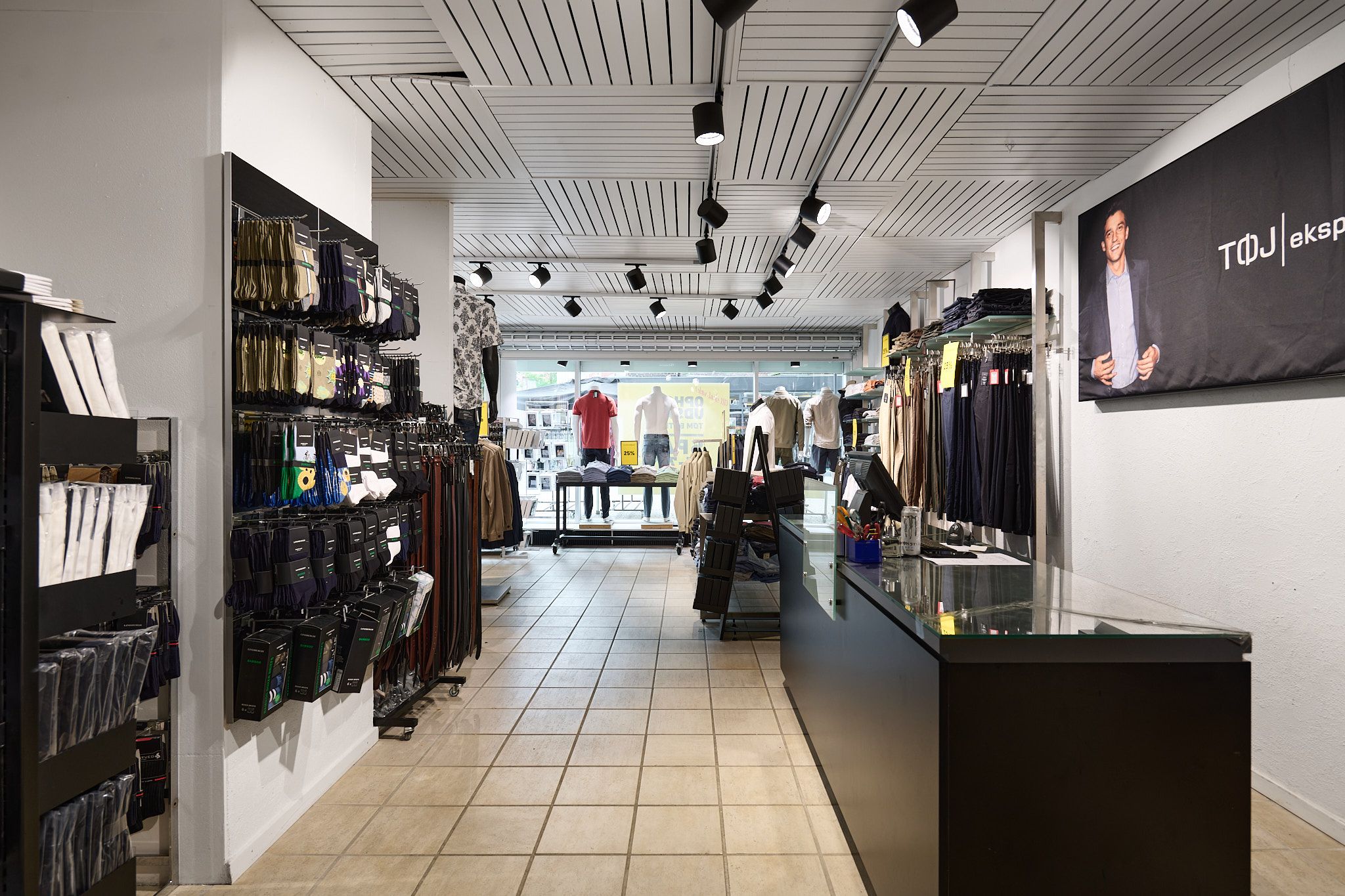 Foto af butik med sort hylde og lysekroner med mænd i tøj på tøjvæg og mand i sort jakke på plakat.