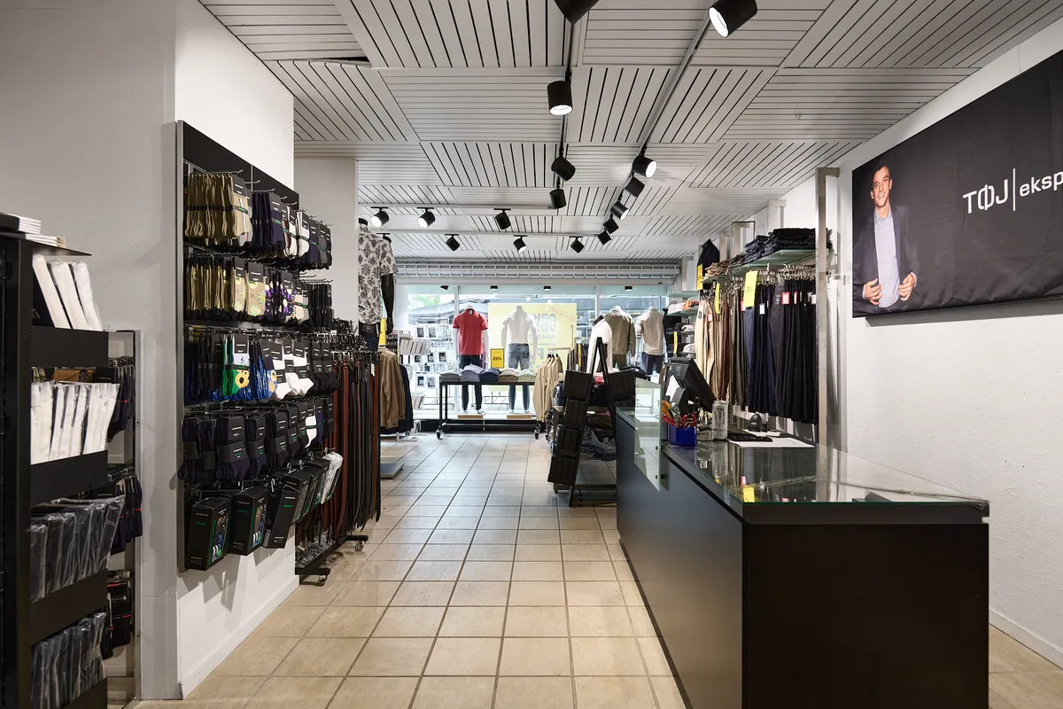 Foto af butik med sort hylde og lysekroner med mænd i tøj på tøjvæg og mand i sort jakke på plakat.