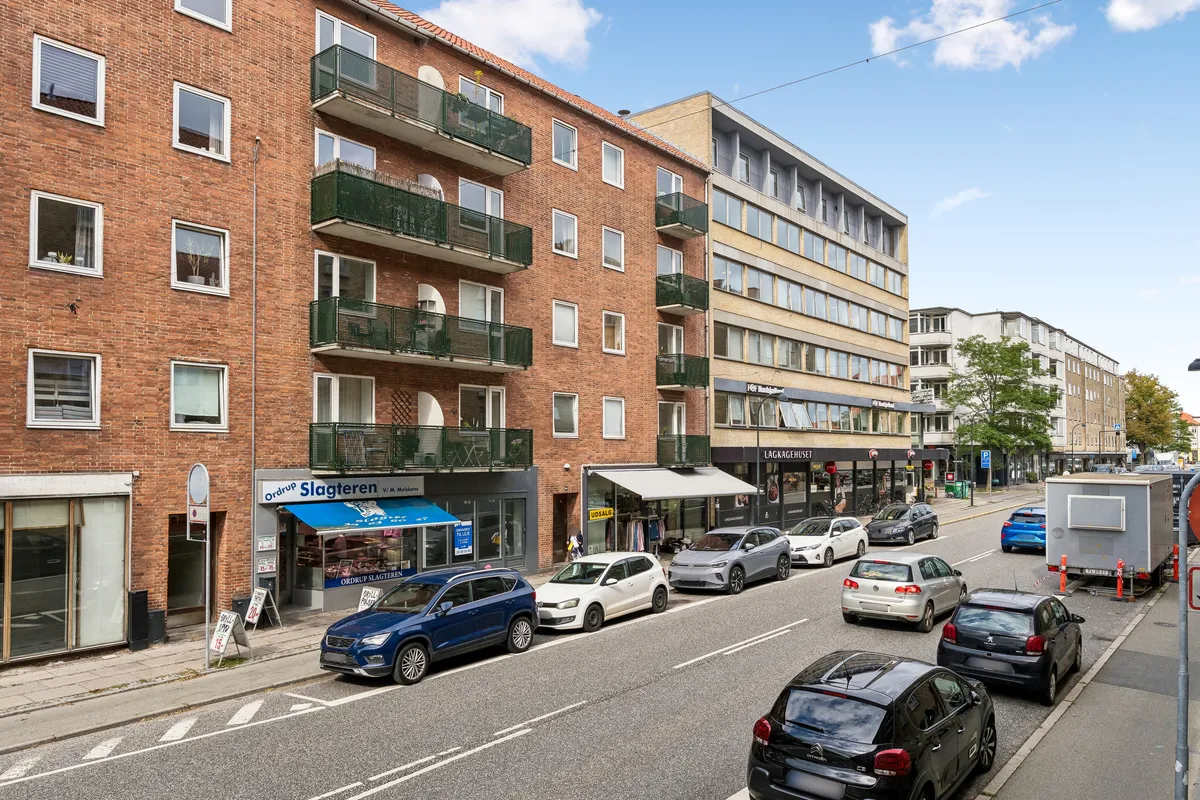 En række forretninger er placeret langs en trafikeret vej, hvor biler er parkeret langs vejkanterne og en stor containervogn venter på en krydsning.
