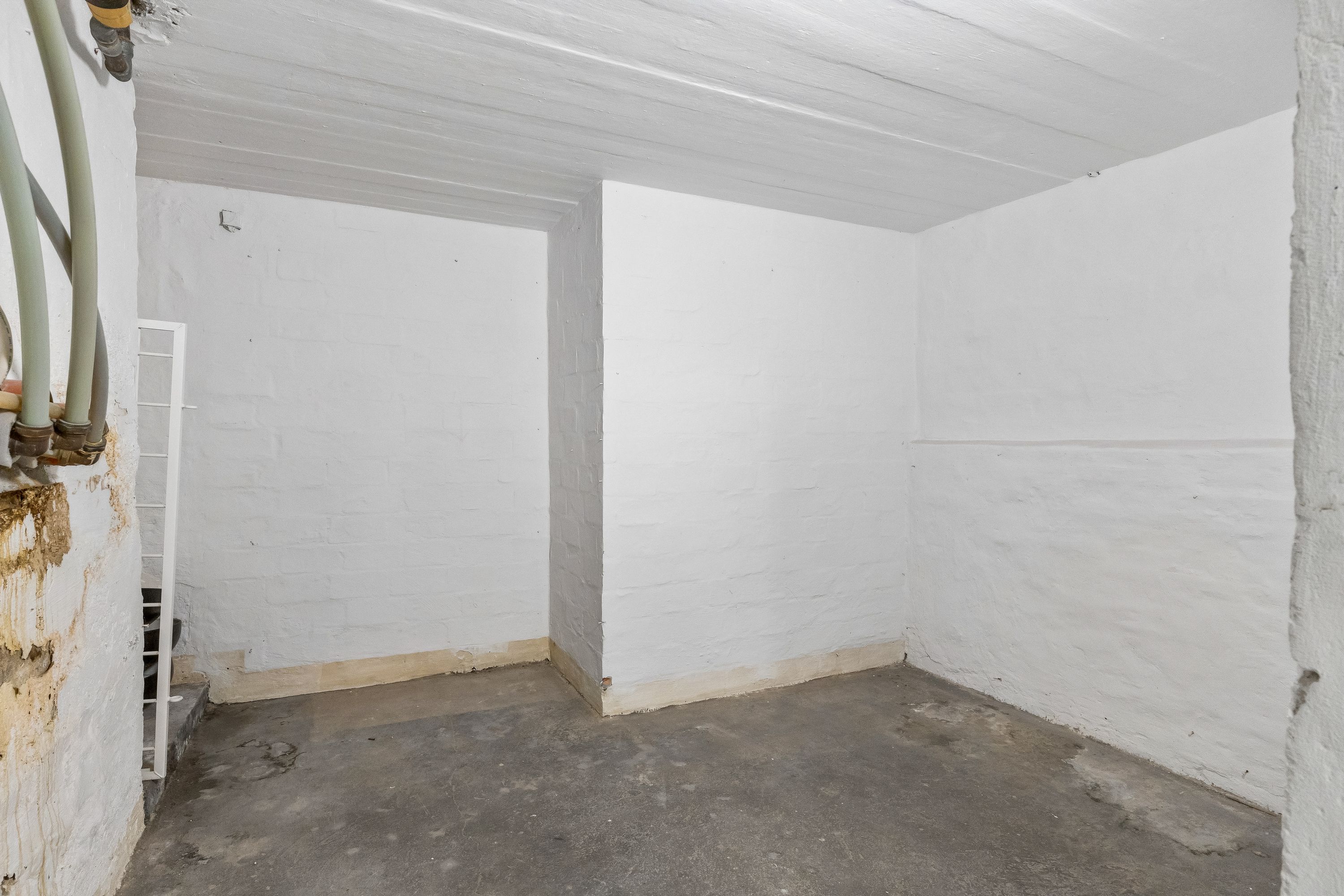 En hvid malet lagerrum med højt og lavt loft, gulv, vægge og røgkanaler. En hvid metaltrappe er fastgjort til væggen.