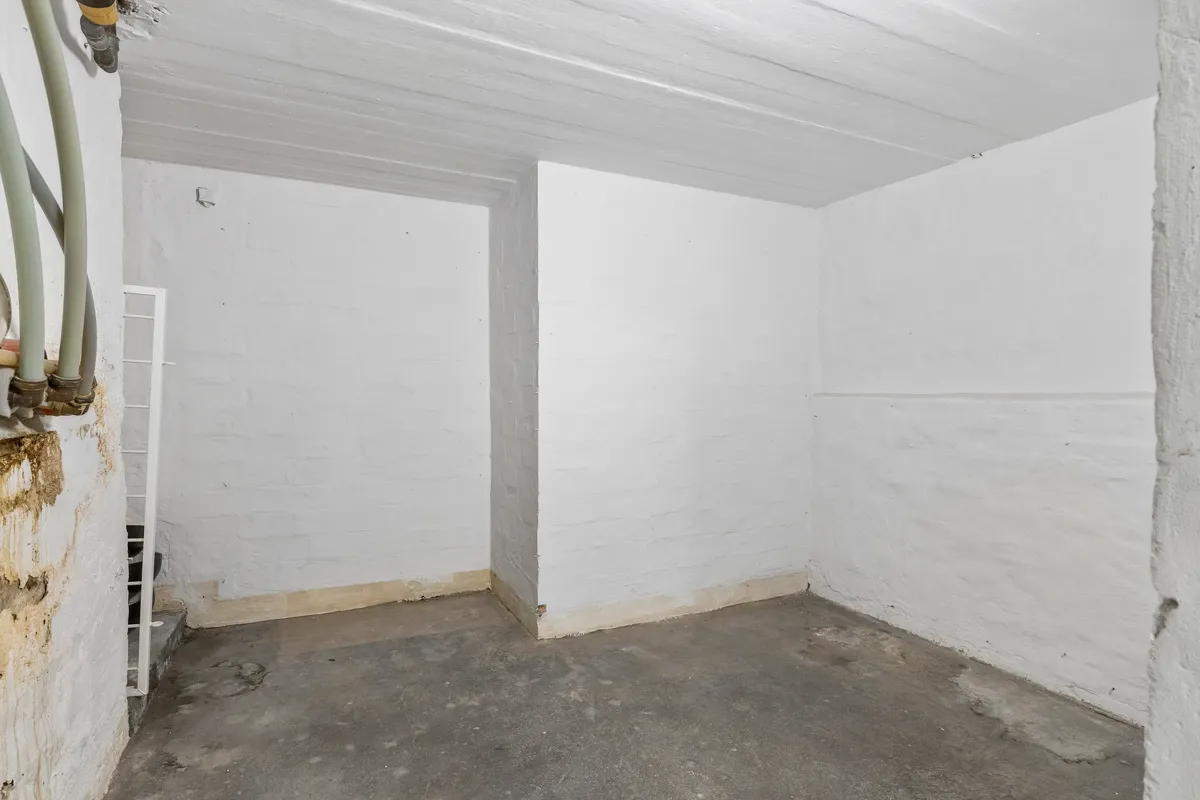 En hvid malet lagerrum med højt og lavt loft, gulv, vægge og røgkanaler. En hvid metaltrappe er fastgjort til væggen.