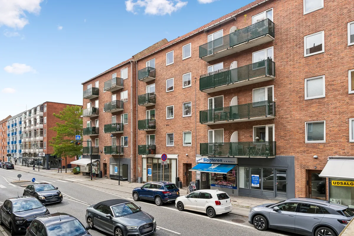 Lejlighedsbygning med udsalgslokaler og parkerede biler på Ordrupvej. Lejlighedsbygningen har flere balkoner og vinduer. Butikkerne har hvide gardiner og gult skilt. Et skilt med blåt stof er op til leje.