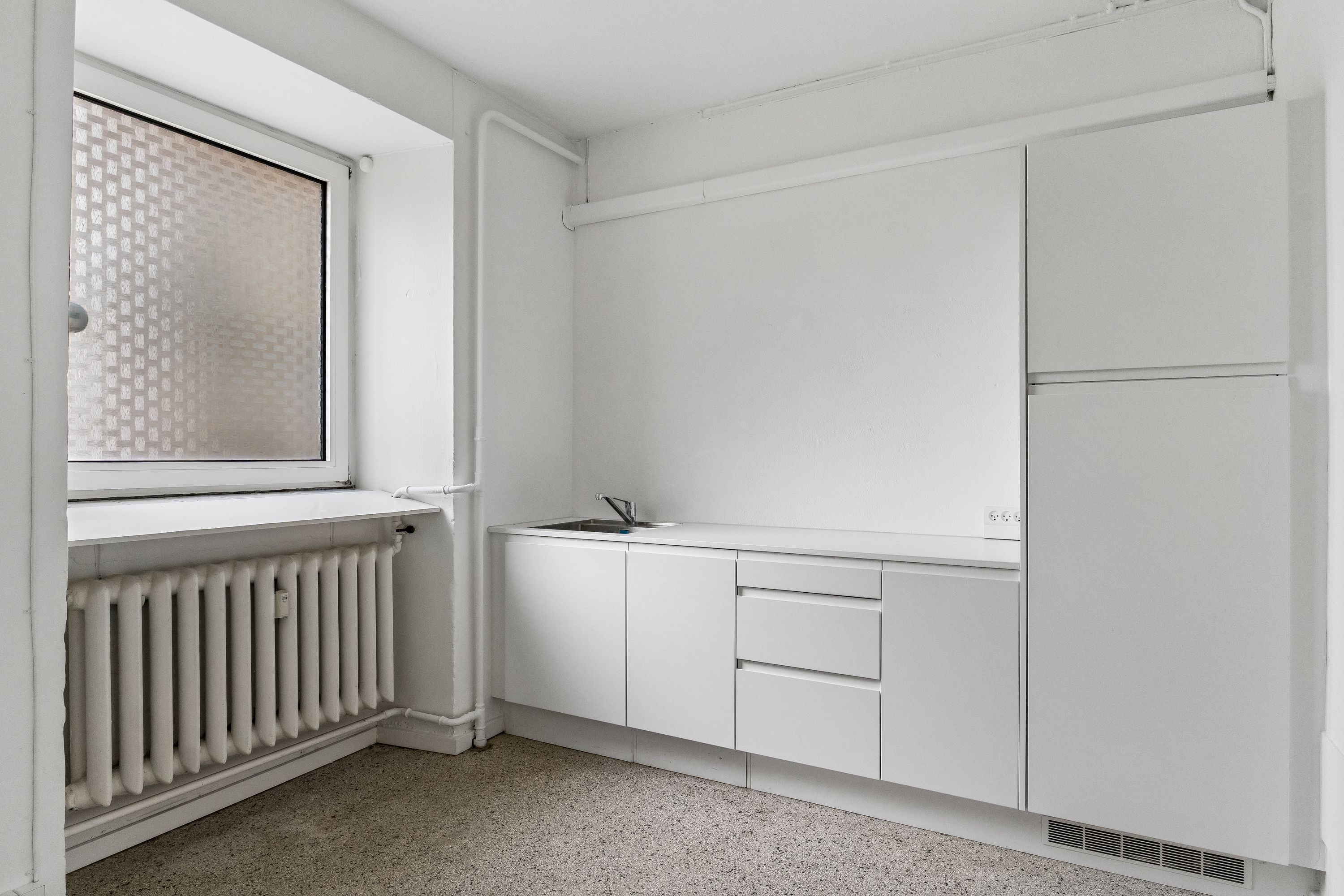 Lejlighed med køkken, radiator og vindueskilt i hvidt og gråt. På gulvet er der granulat.