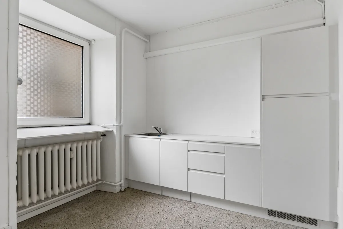 Lejlighed med køkken, radiator og vindueskilt i hvidt og gråt. På gulvet er der granulat.