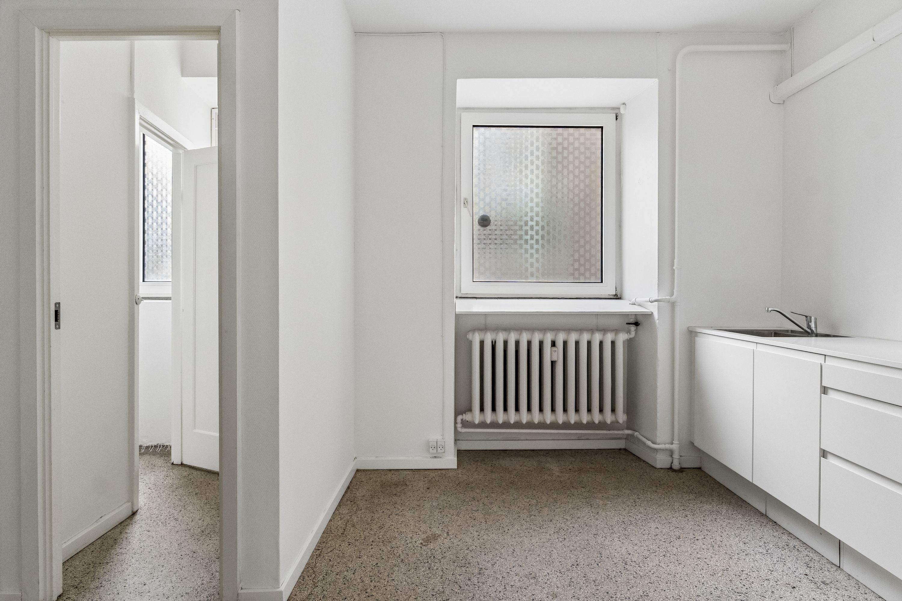 En køkkenrum med hvide vægge, loft, gulv og hvidt køkken med radiator, vandhaner og et rektangulært vindue med glas.