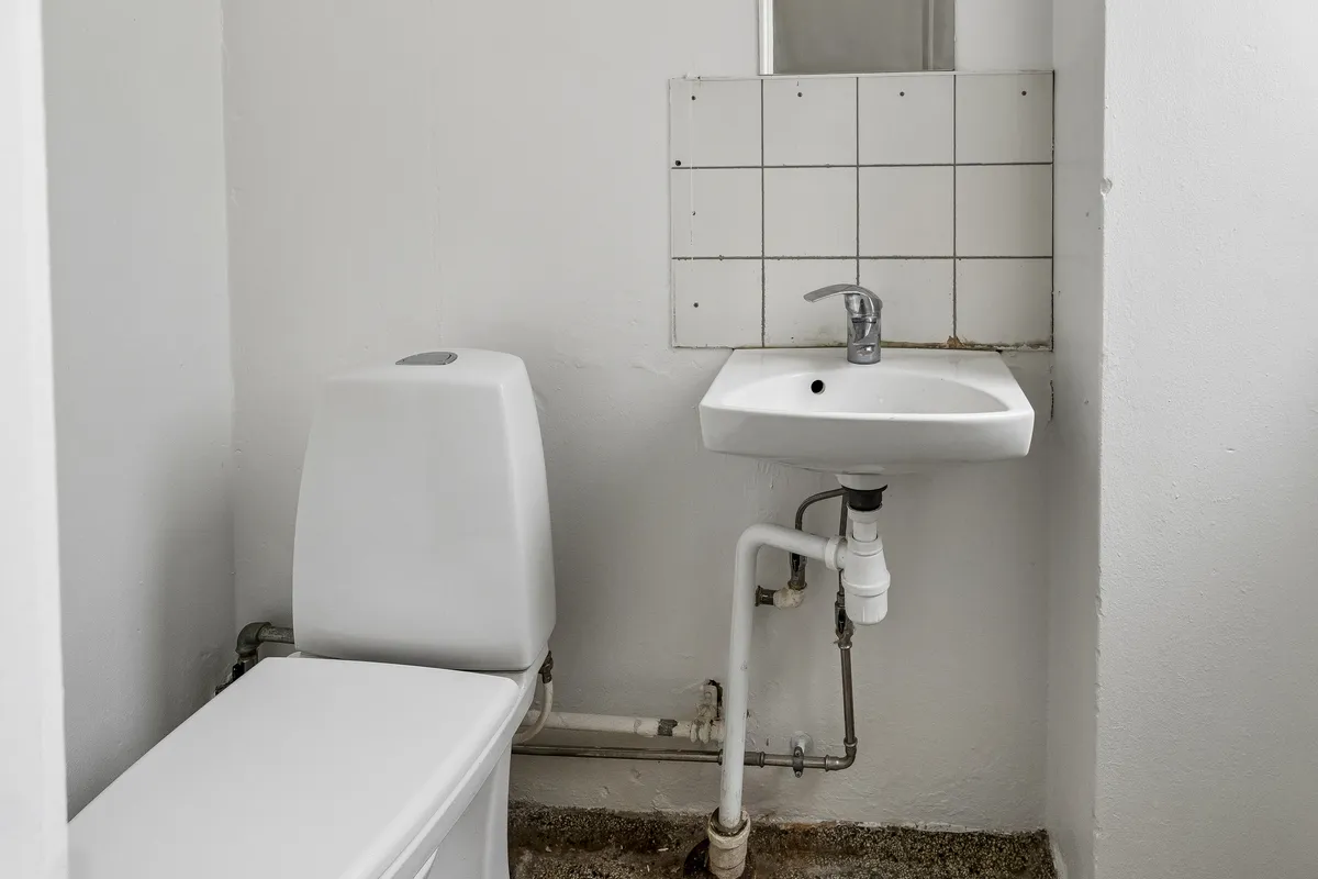 Et lille toilet med hvid toiletbøjle og sænkbar toiletblok samt hvidt vasketøjsbord med håndvask og håndsprøjt