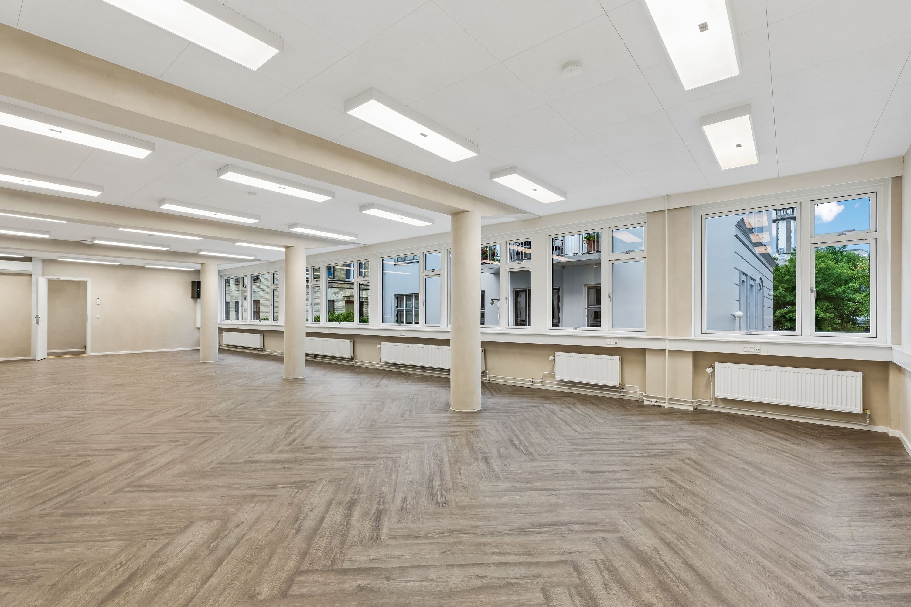 Et tomt kontor med lyse, brede vinduer, hvide loftsbelægninger, moderne lysekroner og parketgulv. Rummet er fuld af naturligt lys, og der er radiatorer installeret langs væggen.