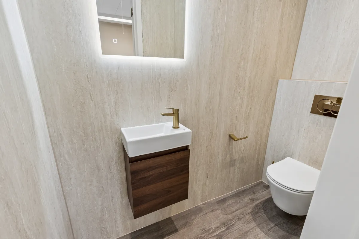 Nyrenoveret og elegant badeværelse med marmor-lignende vægge og en hvid toilet med guldfarvet håndtag. Vandhanen er i guld og der er et lille skab med træfront. Spejlet er indbygget i væggen med lys.