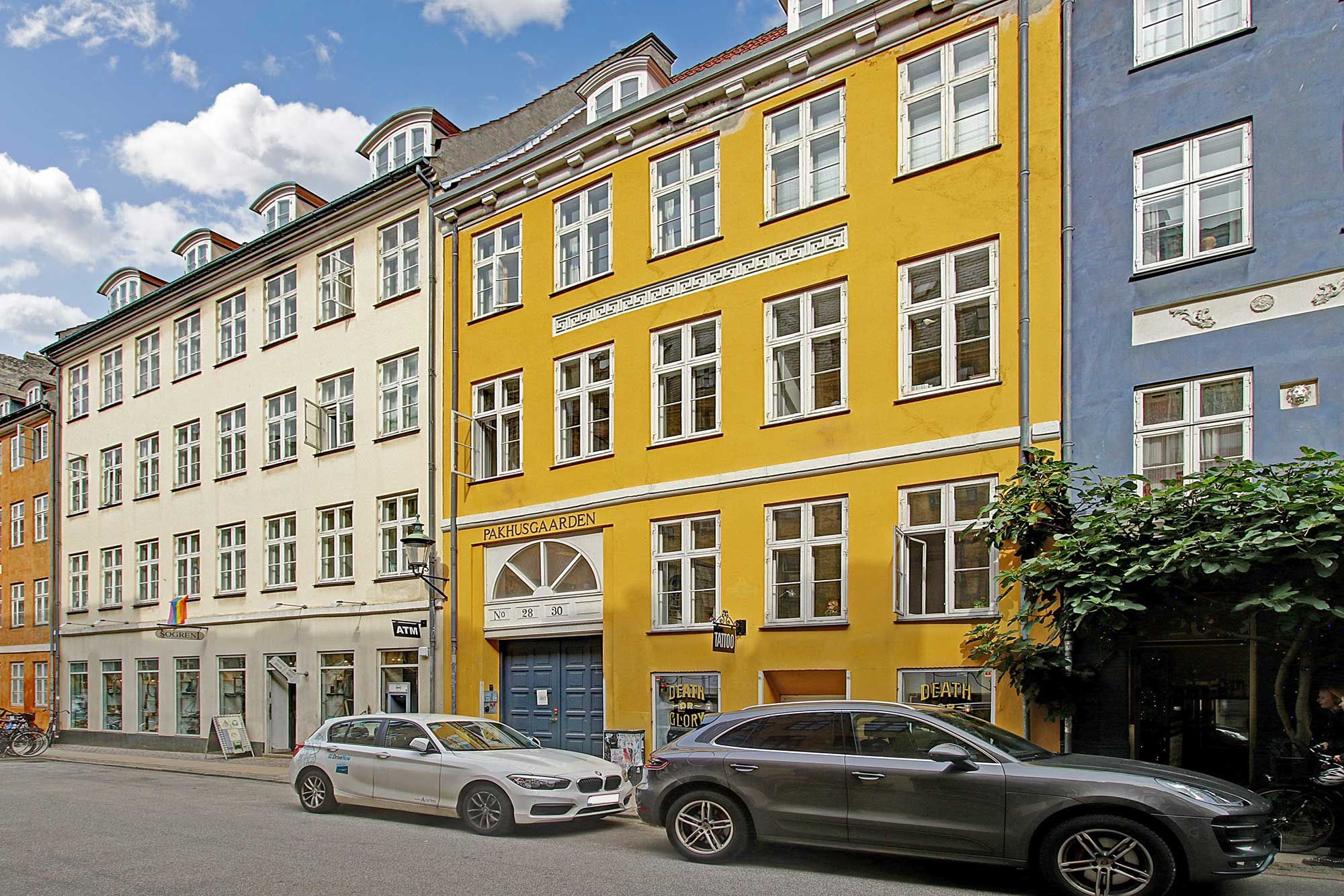 To biler parkeret ved siden af en bygning i Latinerkvarter med røde tag og lysegul facade, hvor der står Pakhusgaarden No 28-30