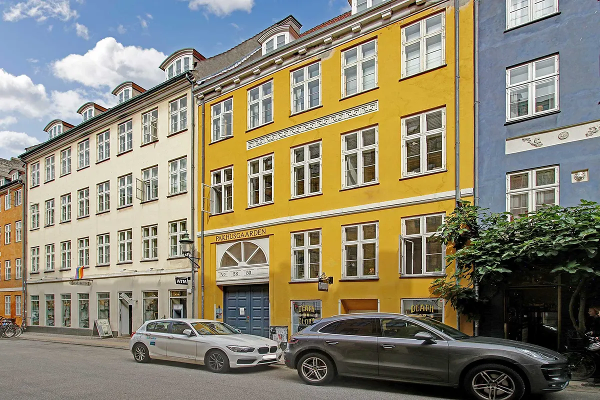 To biler parkeret ved siden af en bygning i Latinerkvarter med røde tag og lysegul facade, hvor der står Pakhusgaarden No 28-30