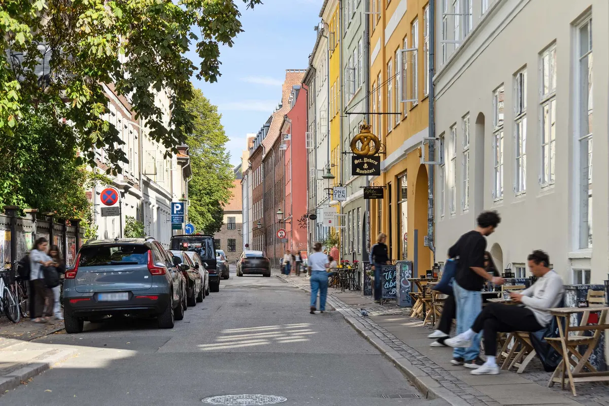 Mange mennesker går på gågaden, og nogle sidder i caféer og spiser. Et gult hus med en skilt med en bagel, og en parkeret bil.