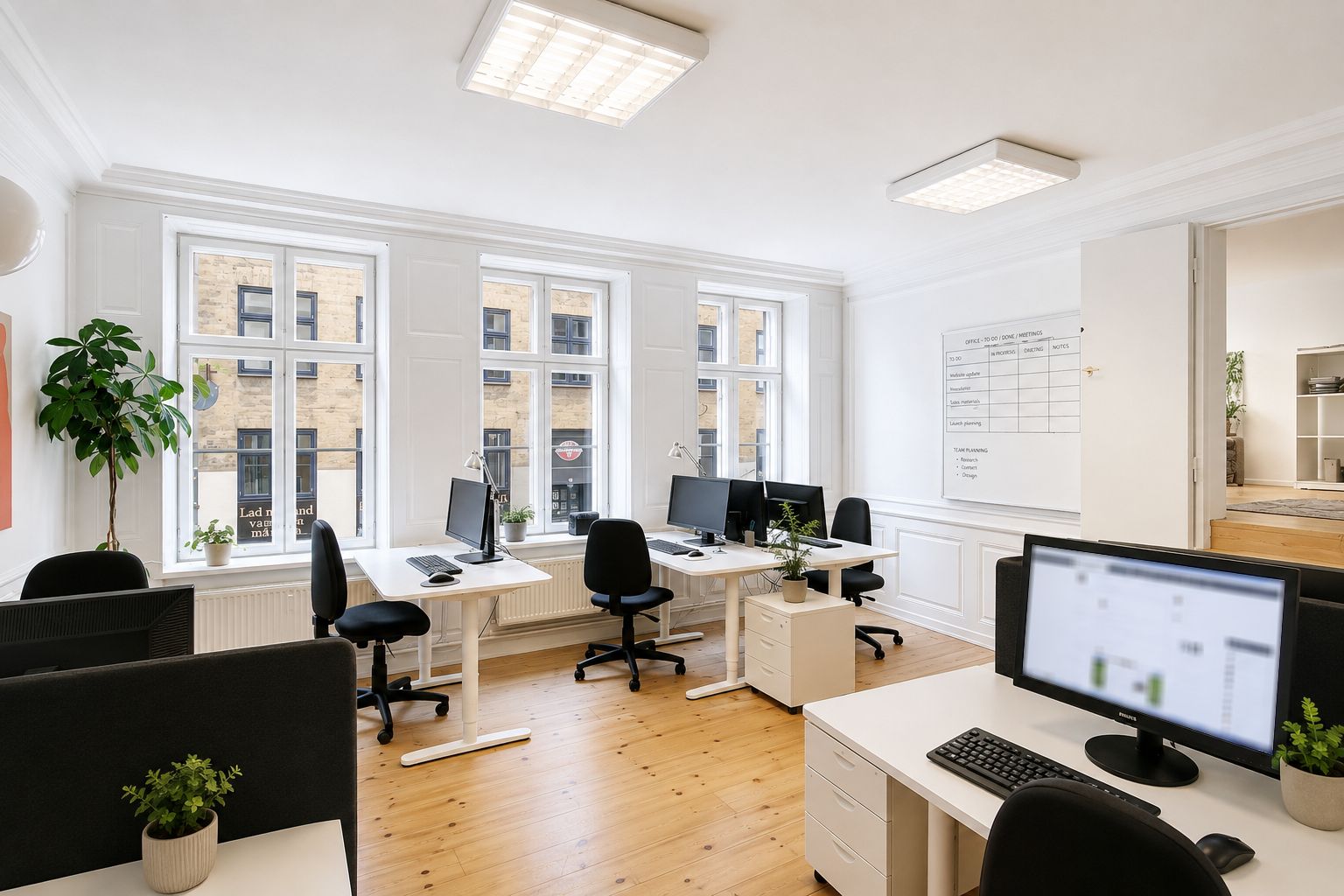 Kontor med højt til loftet, hvide vægge og store vinduer. Der er flere skriveborde med monitorer og computere. En væg er delvist dækket af en tavle med planlægningsnotater.