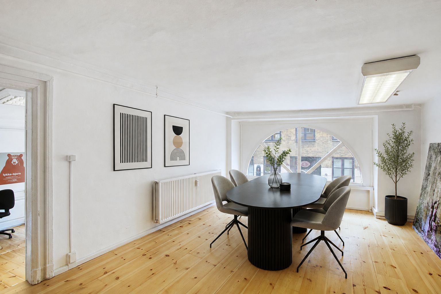 Kontorrum med møbler og lyse, lyse og moderne trægulve og hvide vægge. Runde bord og stole i sort og gråt med grønne planter i hjørner. Arkitektonisk rundt vindue med udsigt til bygninger.