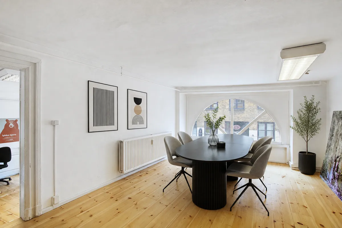Kontorrum med møbler og lyse, lyse og moderne trægulve og hvide vægge. Runde bord og stole i sort og gråt med grønne planter i hjørner. Arkitektonisk rundt vindue med udsigt til bygninger.