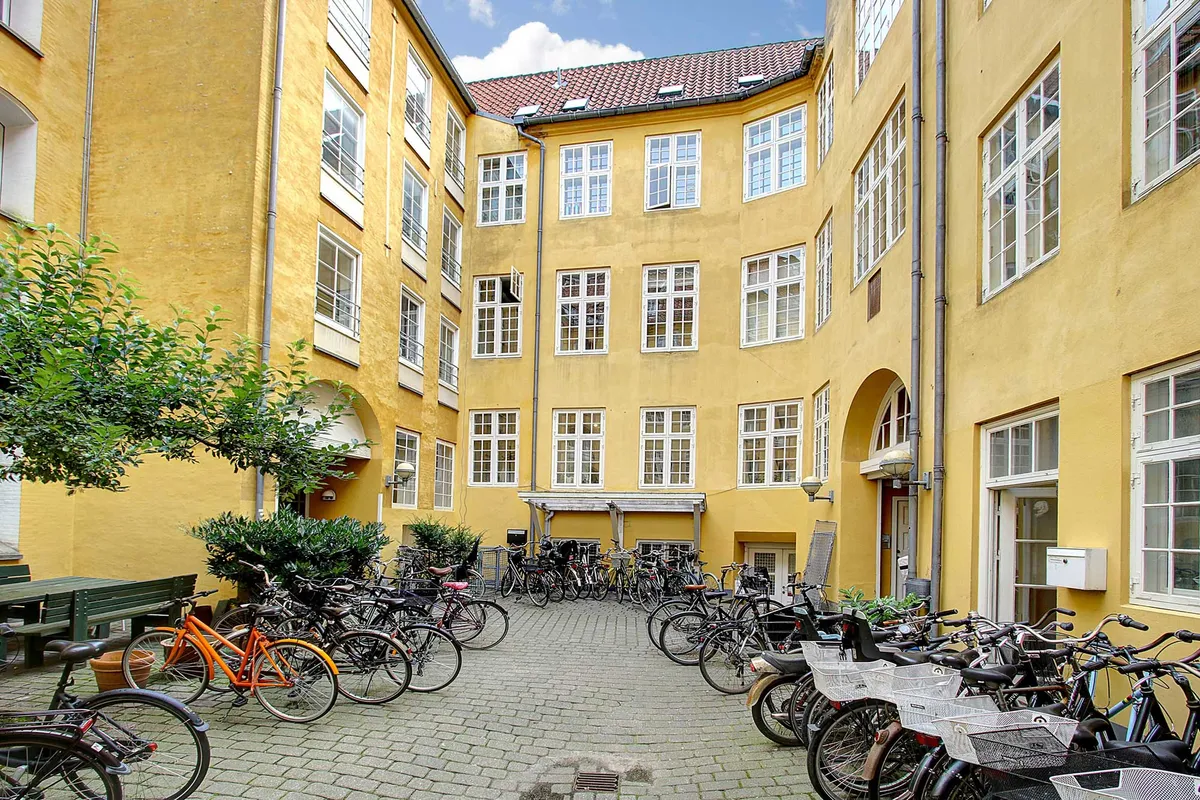 En række cykler parkeret i rækker i et gult bygningskompleks i Latinerkvarteret, ud foran en grøn bænk og en stor træ