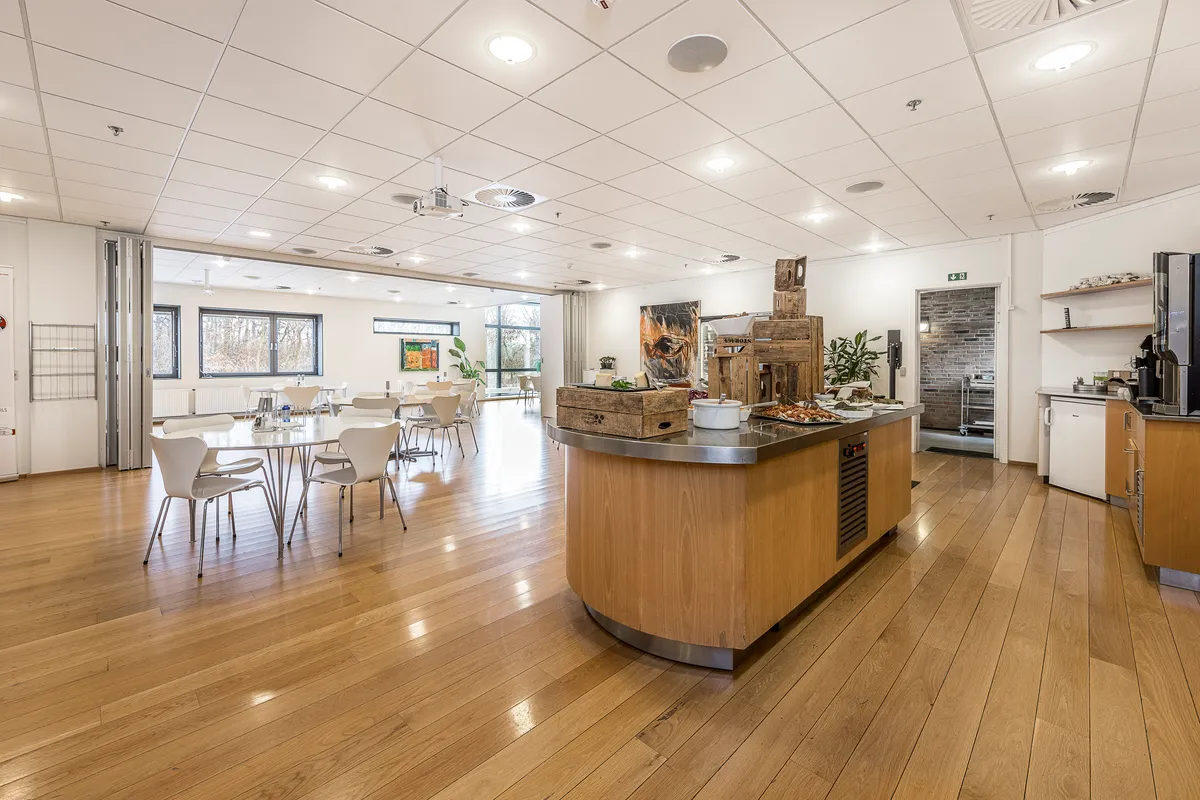 Foto af en stor stue med åben køkkendel og serveringsbord, hvor der er trægulve, lyse lofter, stole og borde samt et køleskab og plante