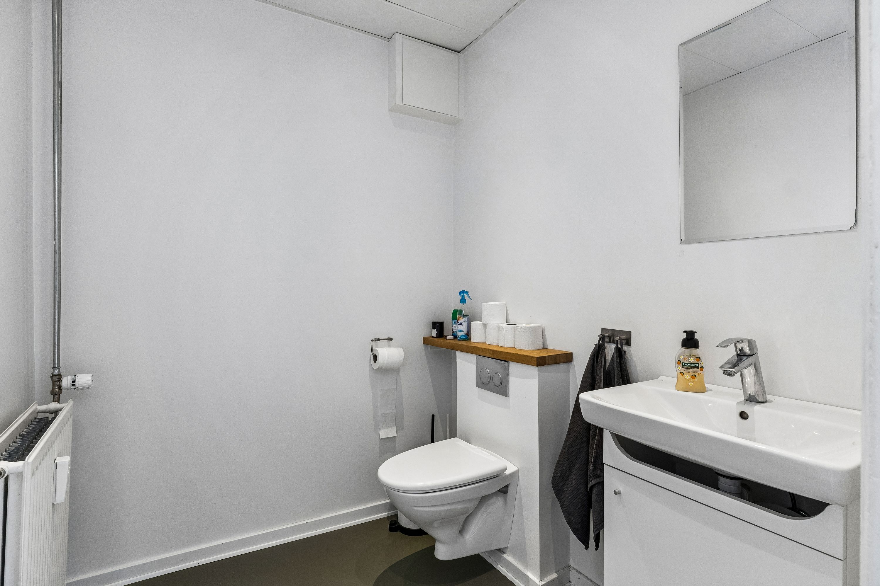 En simpel hvid toilet med toiletpapir og en sæbeflaske på ren hvid vask og et spejl på væggen. Toilettet er i hvidt og har en høj kvalitet.