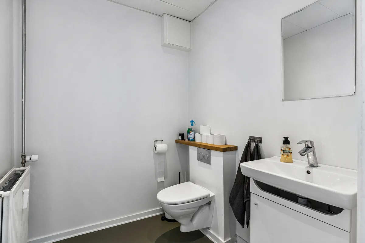 En simpel hvid toilet med toiletpapir og en sæbeflaske på ren hvid vask og et spejl på væggen. Toilettet er i hvidt og har en høj kvalitet.