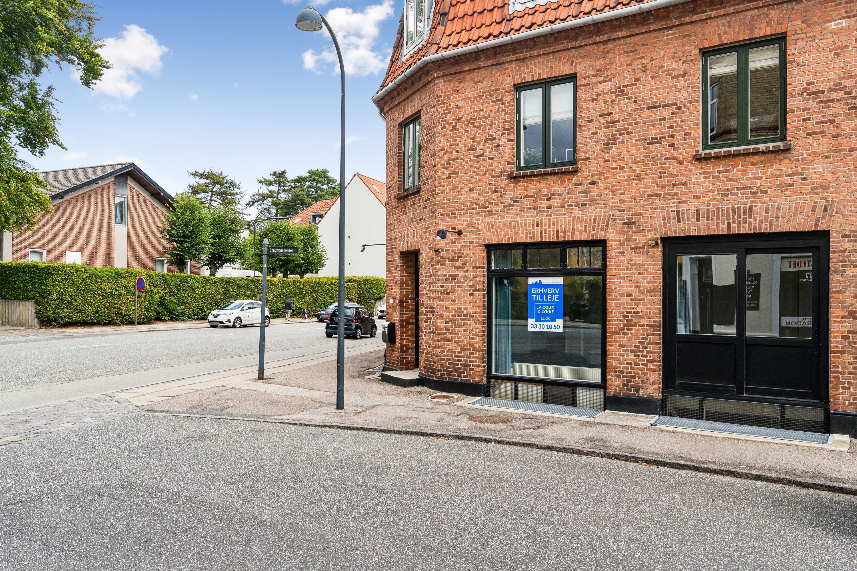 Hjørnebygning med røde mursten og glasruder på Ordrupvej 141 i Charlottenlund. Oplagt til specialbutik eller kontor med store vinduer og glasdøre. Parkerede biler og grønne hække i baggrunden.
