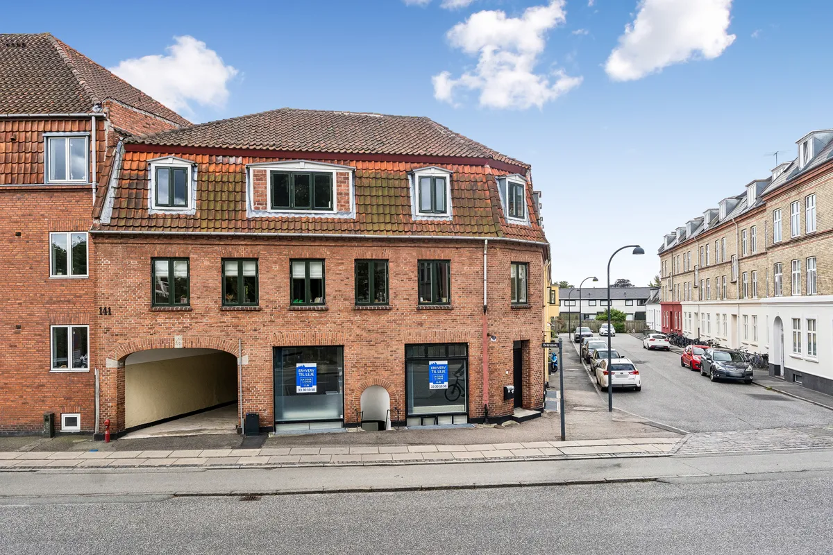 Lejlighed og butik til leje i Charlottenlund. Moderne bygning med røde mursten og store glasvinduer. Parkerede biler og en cykel foran. Solrig dag med lyseregn på himlen