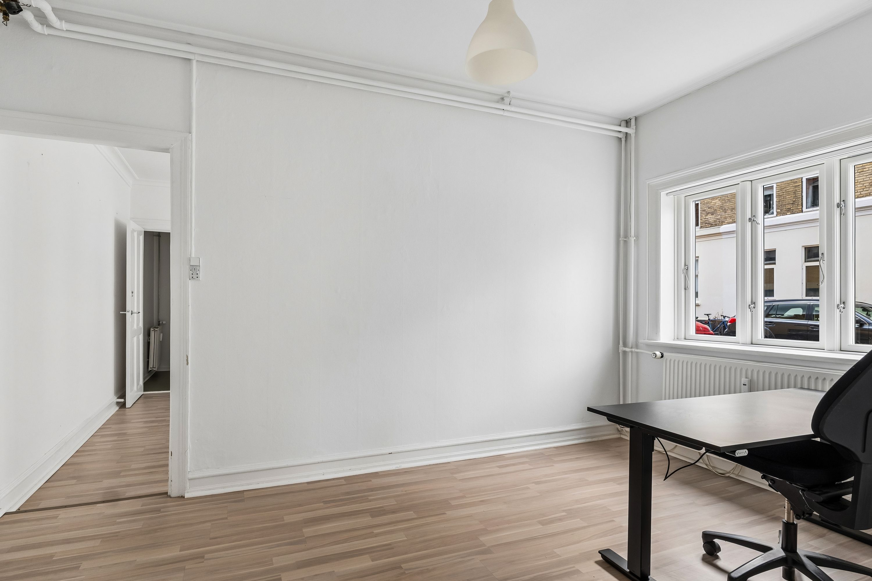 Et rum med hvidt loft og vægge samt hårdt trægulv. Et skrivebord med en sort stol er placeret mod vinduet. Lampen er hængt i loftet. Et andet værelse med åben dør ses i baggrunden.