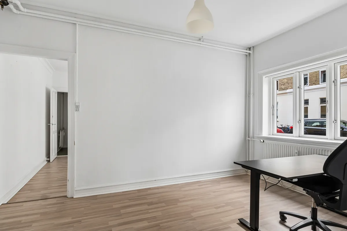 Et rum med hvidt loft og vægge samt hårdt trægulv. Et skrivebord med en sort stol er placeret mod vinduet. Lampen er hængt i loftet. Et andet værelse med åben dør ses i baggrunden.